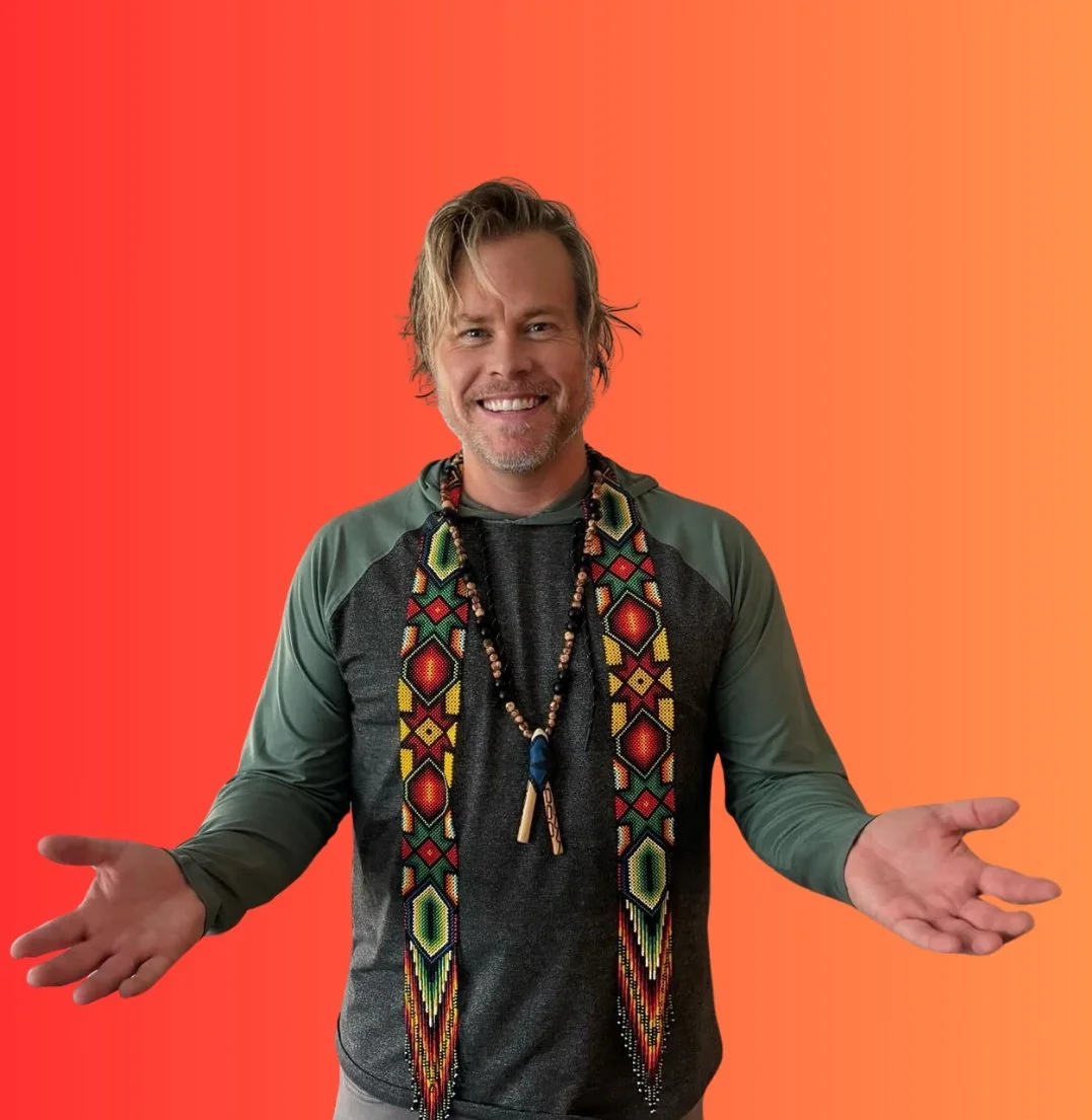 A smiling man with light hair and a beard, wearing a dark jacket with green sleeves, and beaded necklaces, standing with his arms outstretched against a gradient orange background.