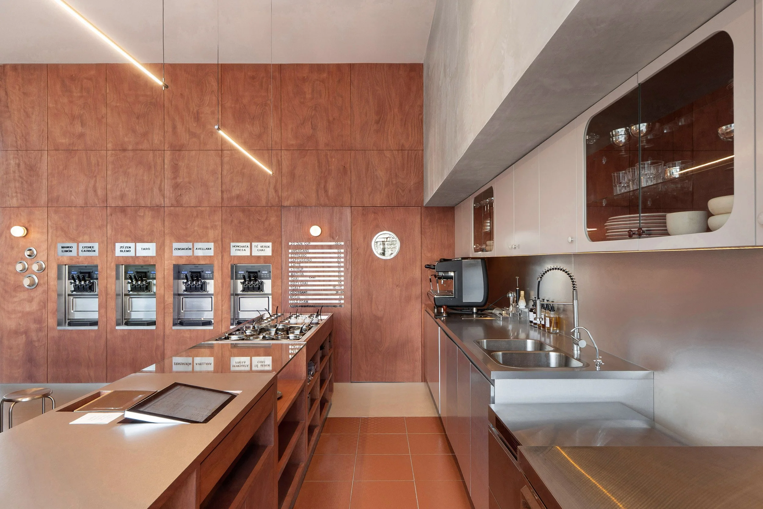 Interior design of Zo'Zen in San Pedro Garza García by Mariel Lozano Studio, showing frozen yogurt dispensers set in warm teak paneling alongside a sleek stainless steel topping counter display.
