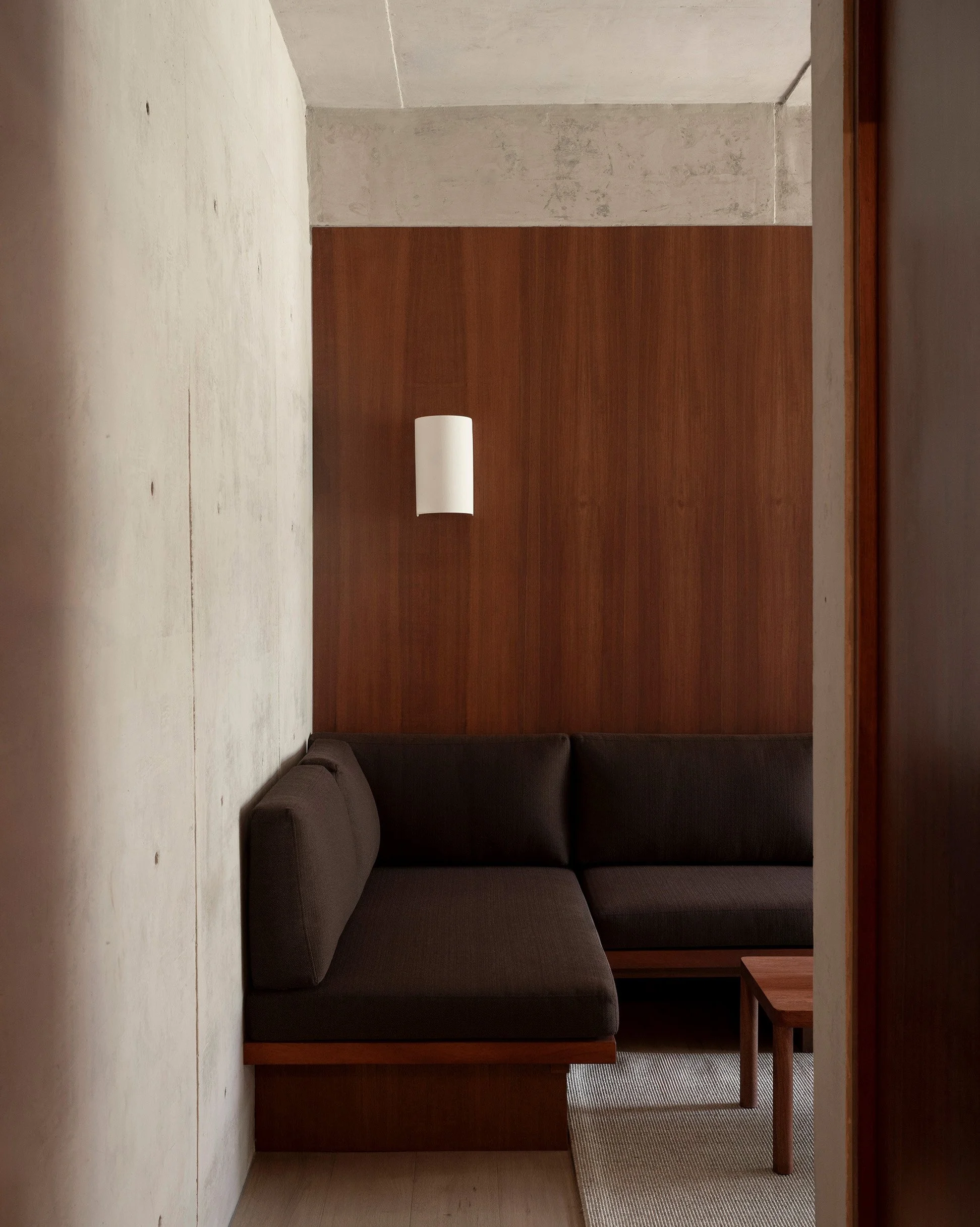 Custom built-in L-shaped sofa set against rich wood wall paneling and raw concrete at San Luis hospitality apartment in Mexico City, interior design by Mariel Lozano Studio, photo by Fabian Martinez.