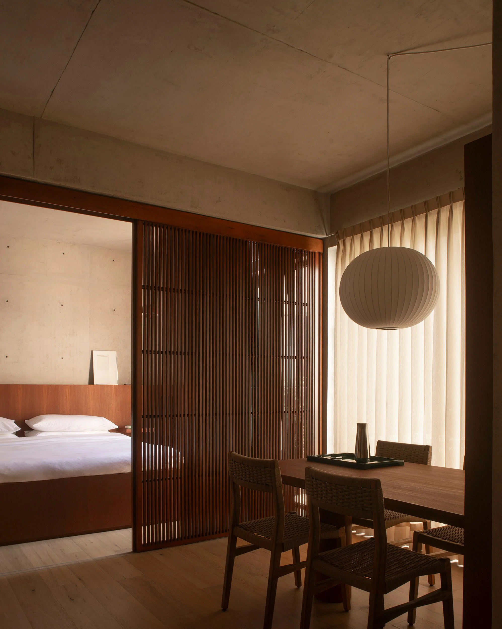 Open-plan layout showing the dining area with a paper pendant lamp opening into the bedroom through timber sliding screens at San Luis apartment in Mexico City by Mariel Lozano Studio, photo by Fabian Martinez.