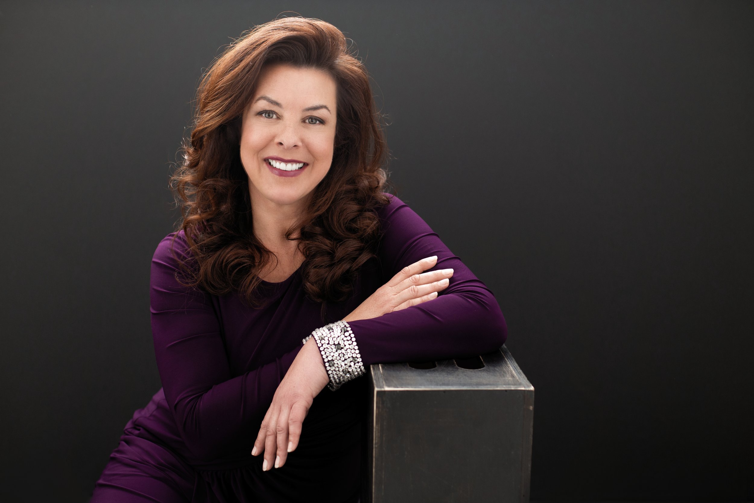 Smiling woman with brown curly hair, wearing a purple dress and silver bracelet, against a dark background.