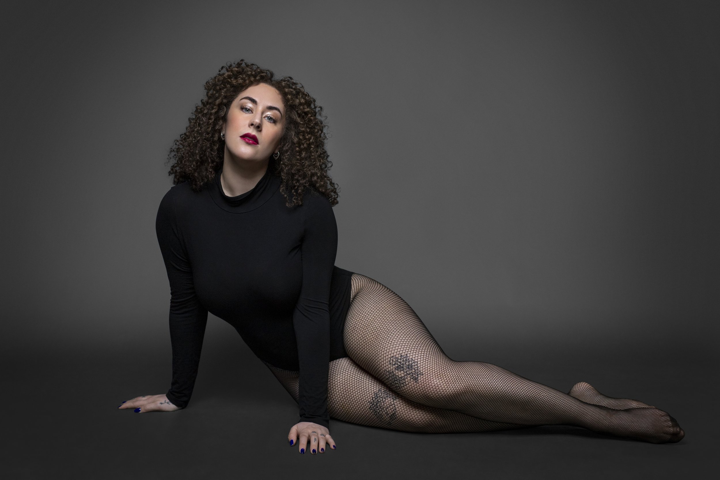 Woman with curly hair in a black bodysuit and fishnet stockings poses against a gray background.