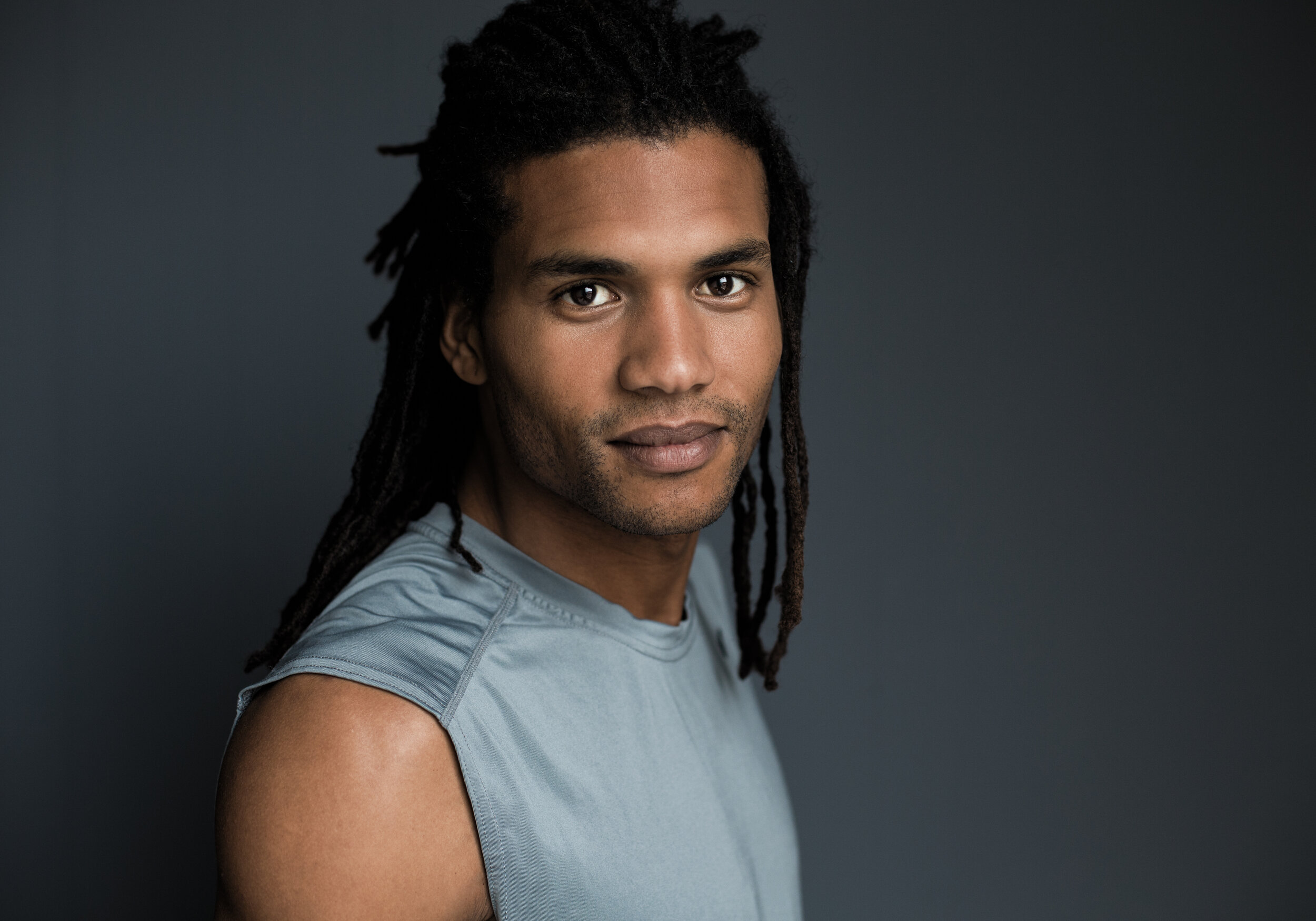 Portrait of a person with long hair wearing a sleeveless shirt against a dark background.