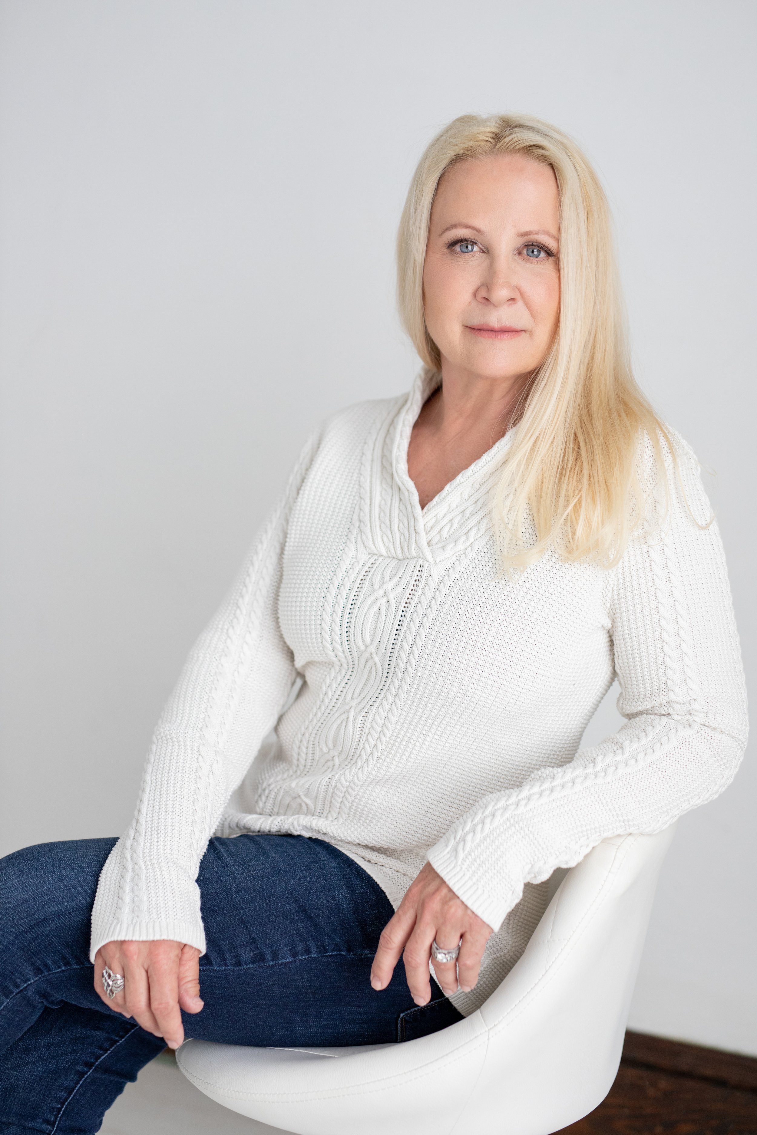 A woman with long blonde hair seated on a white chair, wearing a white knitted sweater and blue jeans.