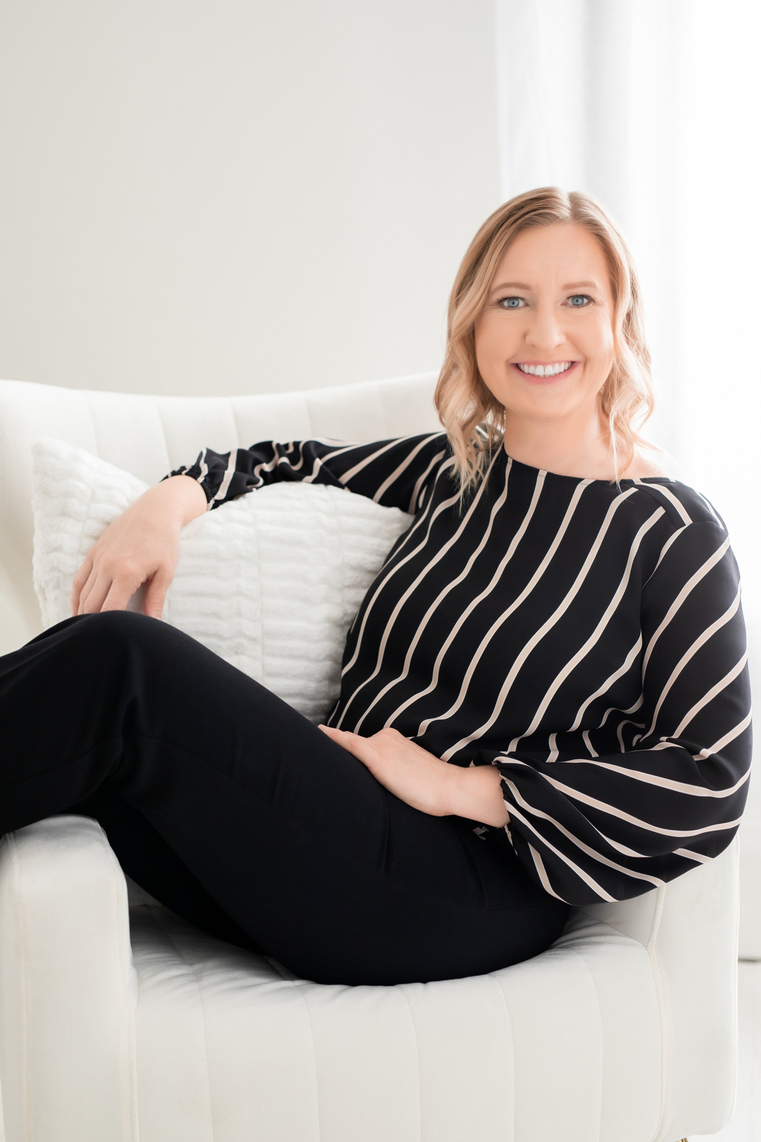 Smiling woman with blonde hair sitting in a white chair, wearing a black and white striped blouse and black pants.