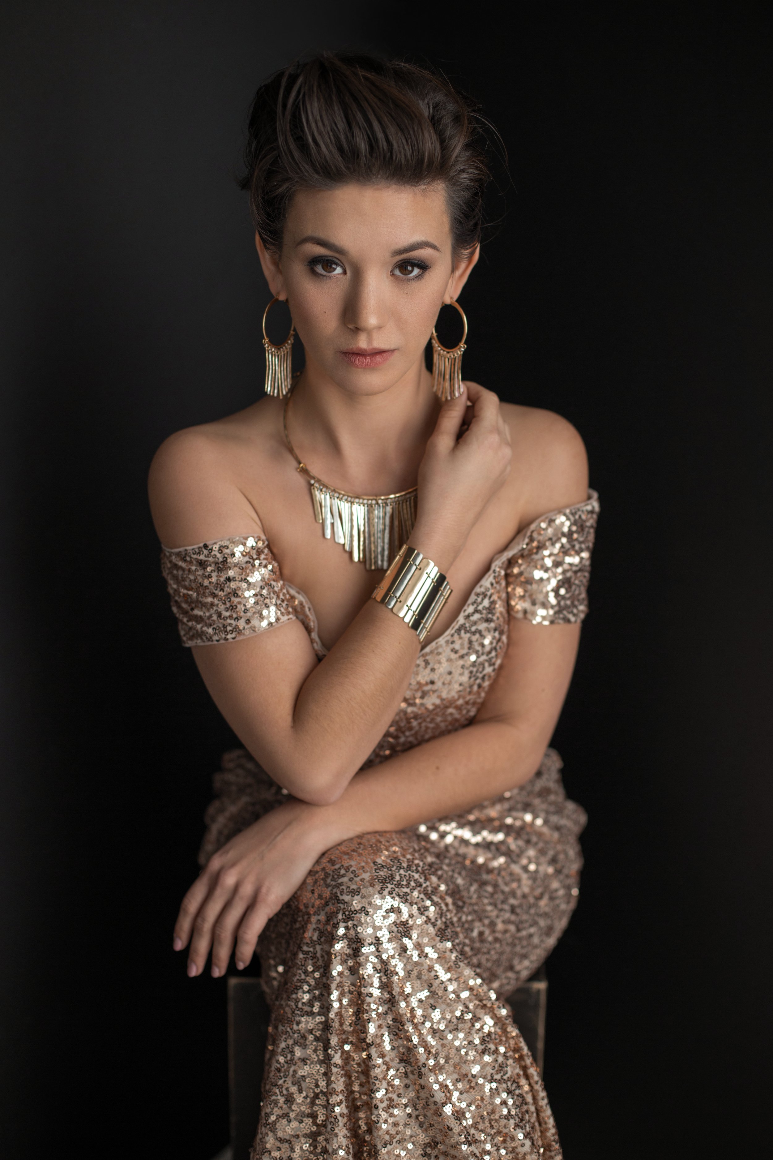 Woman wearing a gold sequin dress, gold hoop earrings, and matching bracelet and necklace against a black background.
