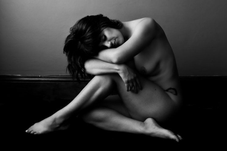 Black and white photograph of a nude woman sitting on the floor, hugging her knees with her head resting on her arms.