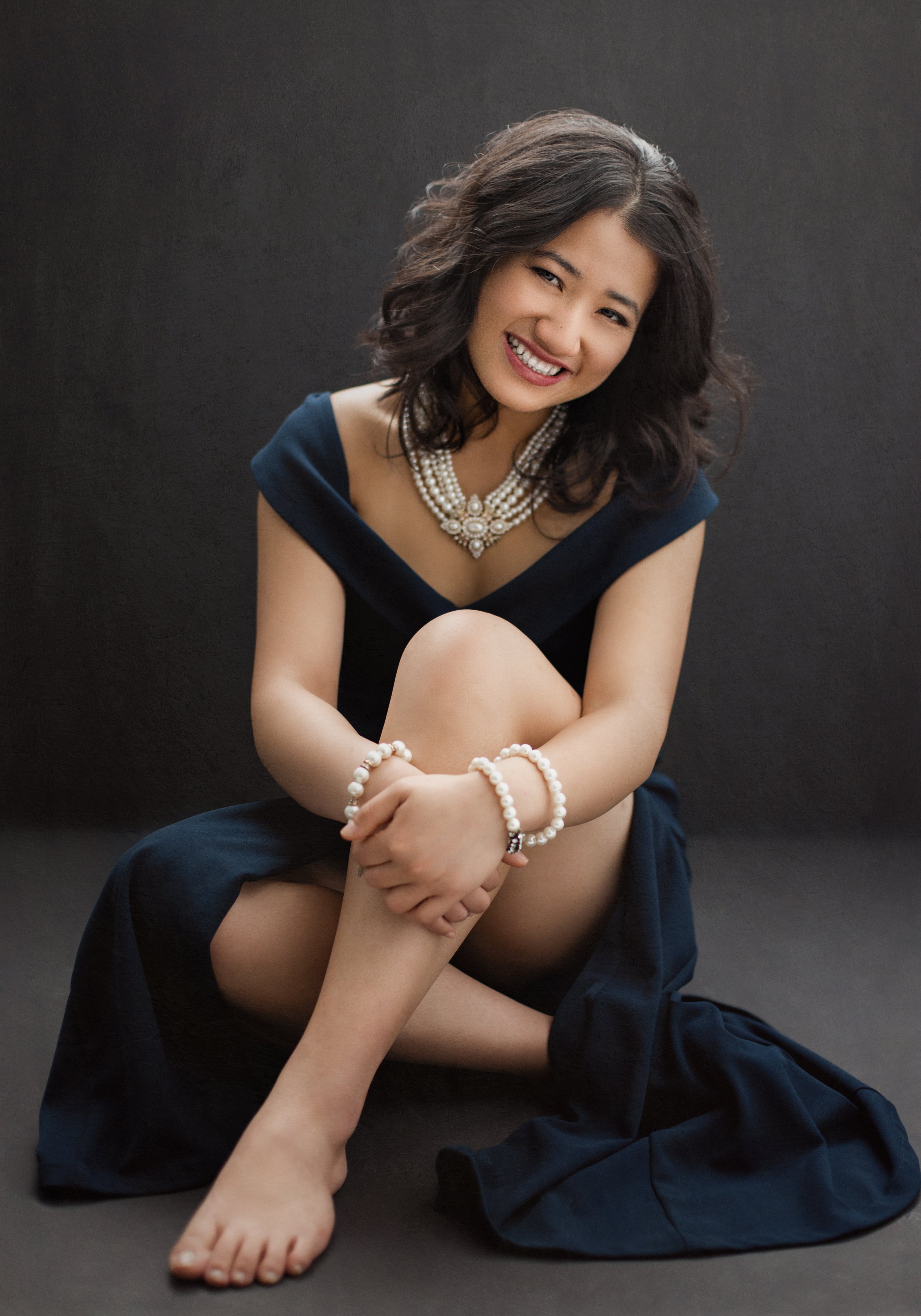 Smiling woman sitting in a navy blue dress with pearl jewelry.