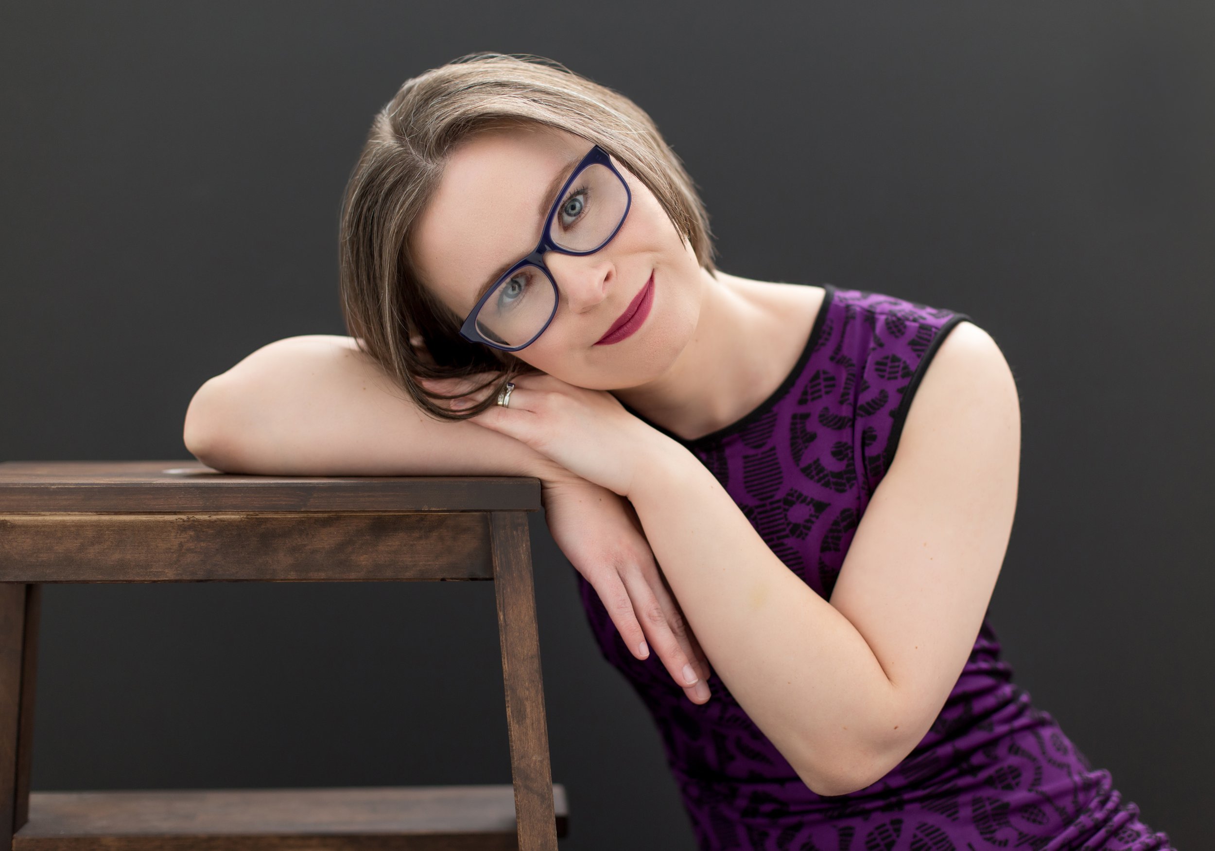 Woman with short hair and glasses leaning on a wooden chair, wearing a purple patterned dress, smiling at the camera with a neutral background.