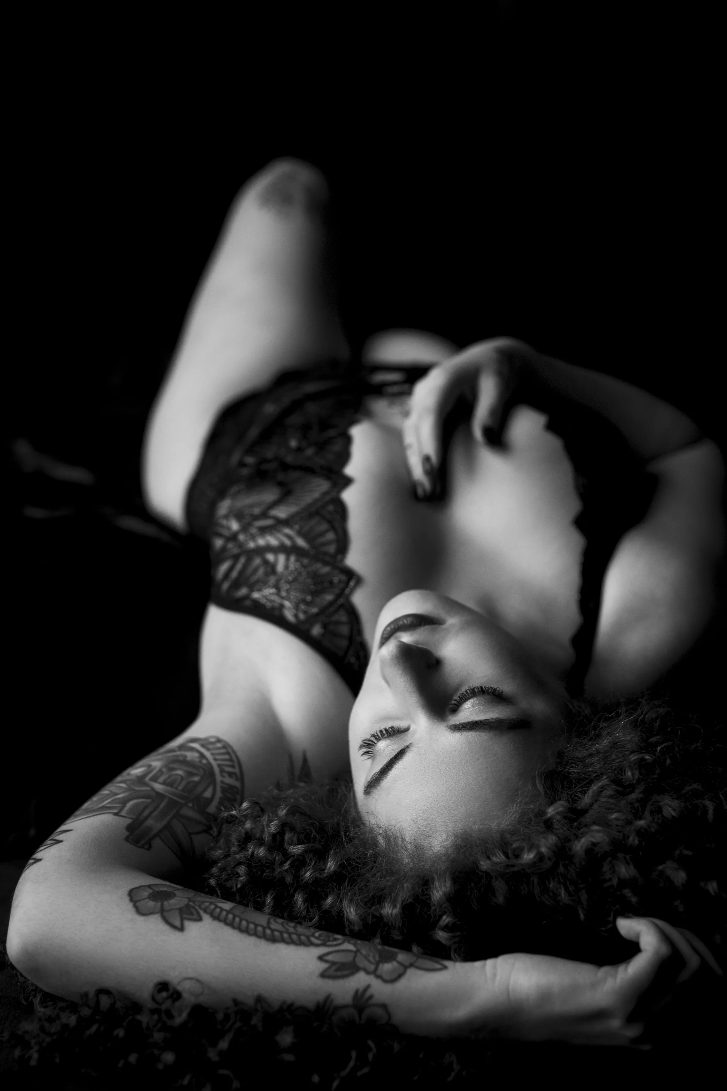 Black and white photo of a person lying down, wearing lace lingerie, with tattoos on their arm, eyes closed.