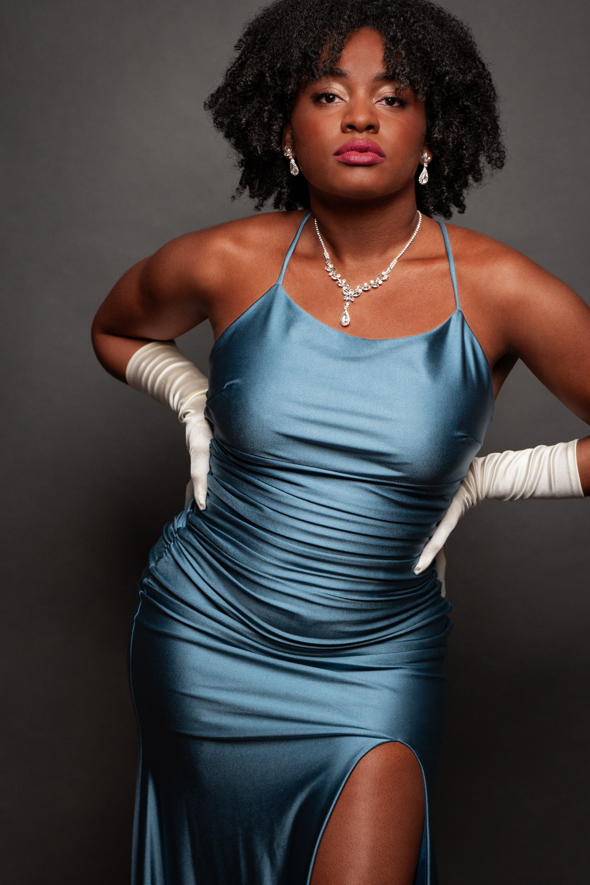 Person wearing a blue satin evening gown with a thigh-high slit, white gloves, silver jewelry, and curly hair.