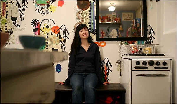 A woman with black hair sitting on a dark wooden bench in a kitchen, wearing a black shirt and jeans, with a colorful mural and a mirror on the wall behind her, and a white stove nearby.
