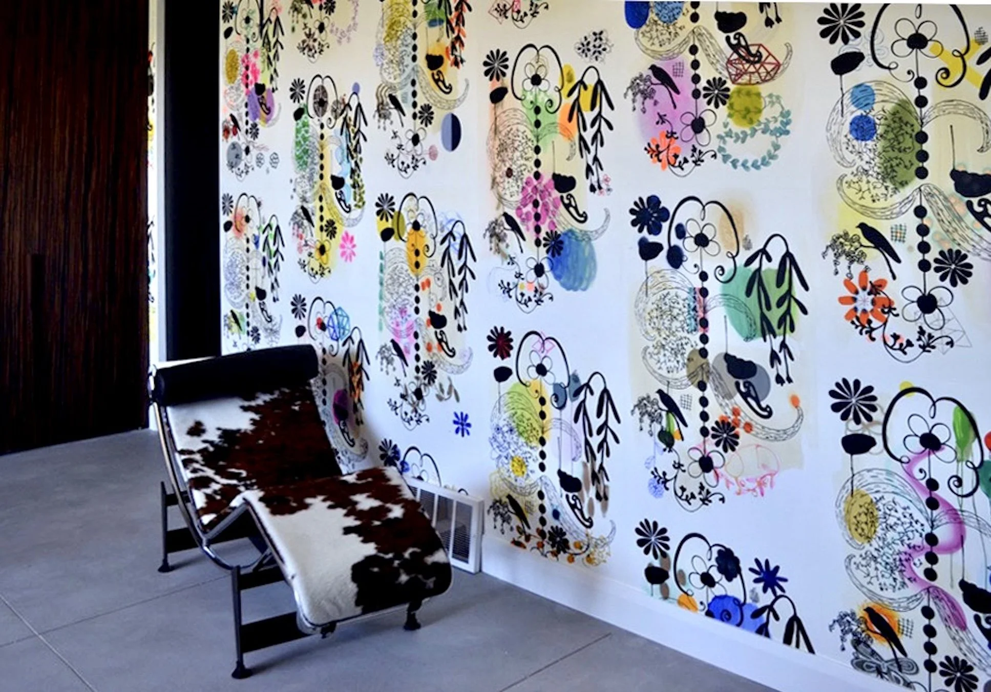 An interior space with a white wall decorated with colorful, abstract, floral, and geometric patterns. On the left side, there is a dark wooden wall and two chairs with cowhide upholstery placed against the white wall. The floor is gray, and a small 