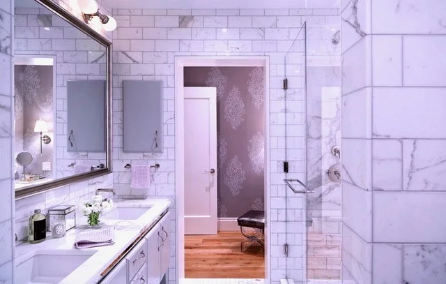 A modern bathroom with white marble tiles, a double sink vanity with a mirror, a small potted plant, and a glass shower enclosure. There is a door at the back leading to another room.