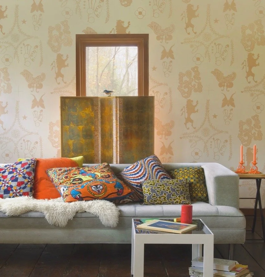 Living room with patterned wallpaper, a white sofa covered with colorful pillows, a sheepskin throw, a small side table with pink candles, and a white coffee table with books, a red candle, and a red cup. A window with a view of trees and a metal she