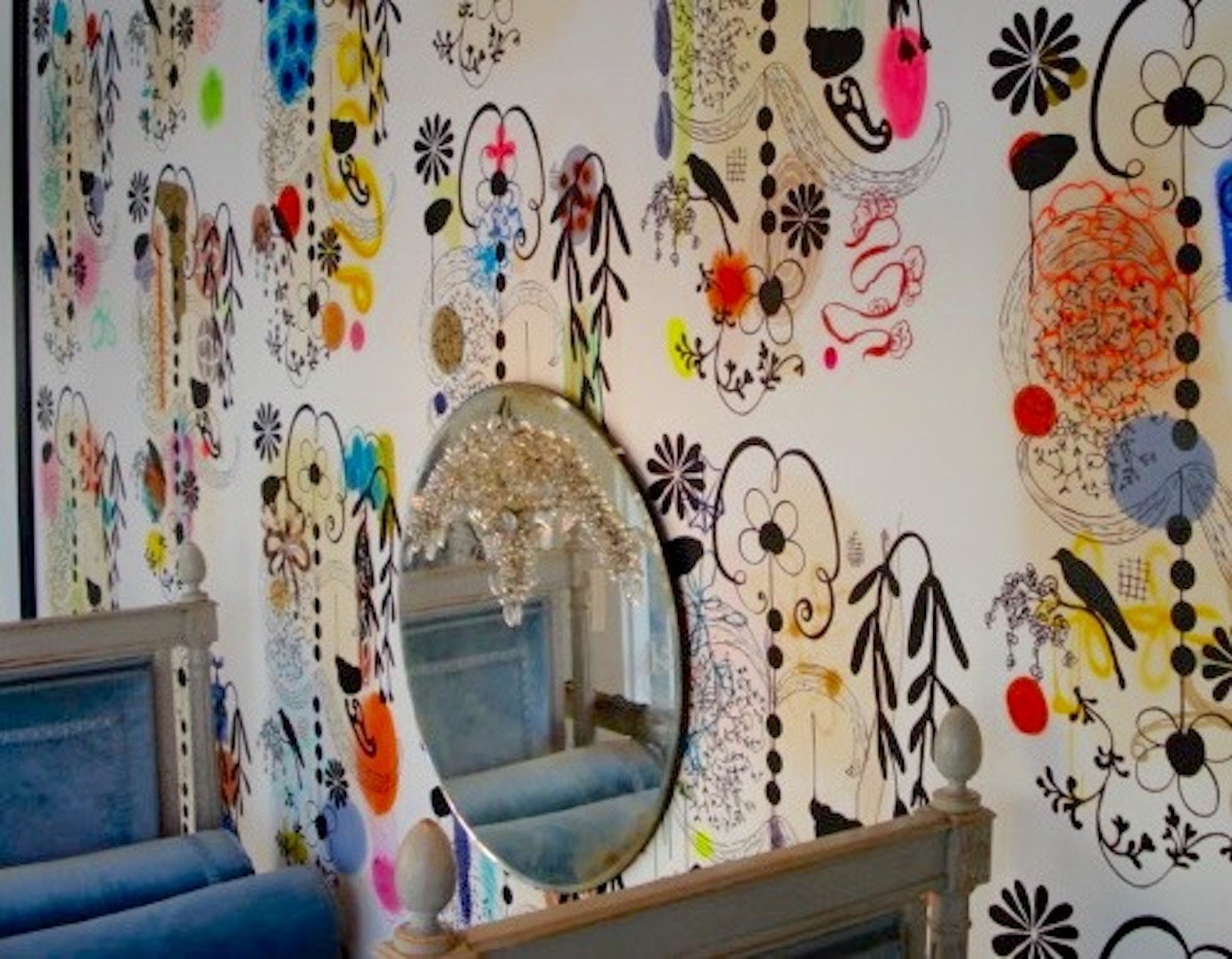 A bedroom with a white wall decorated with colorful and black abstract floral and leaf patterns, a large oval mirror, a wooden bed with a blue headboard, and a chandelier reflected in the mirror.