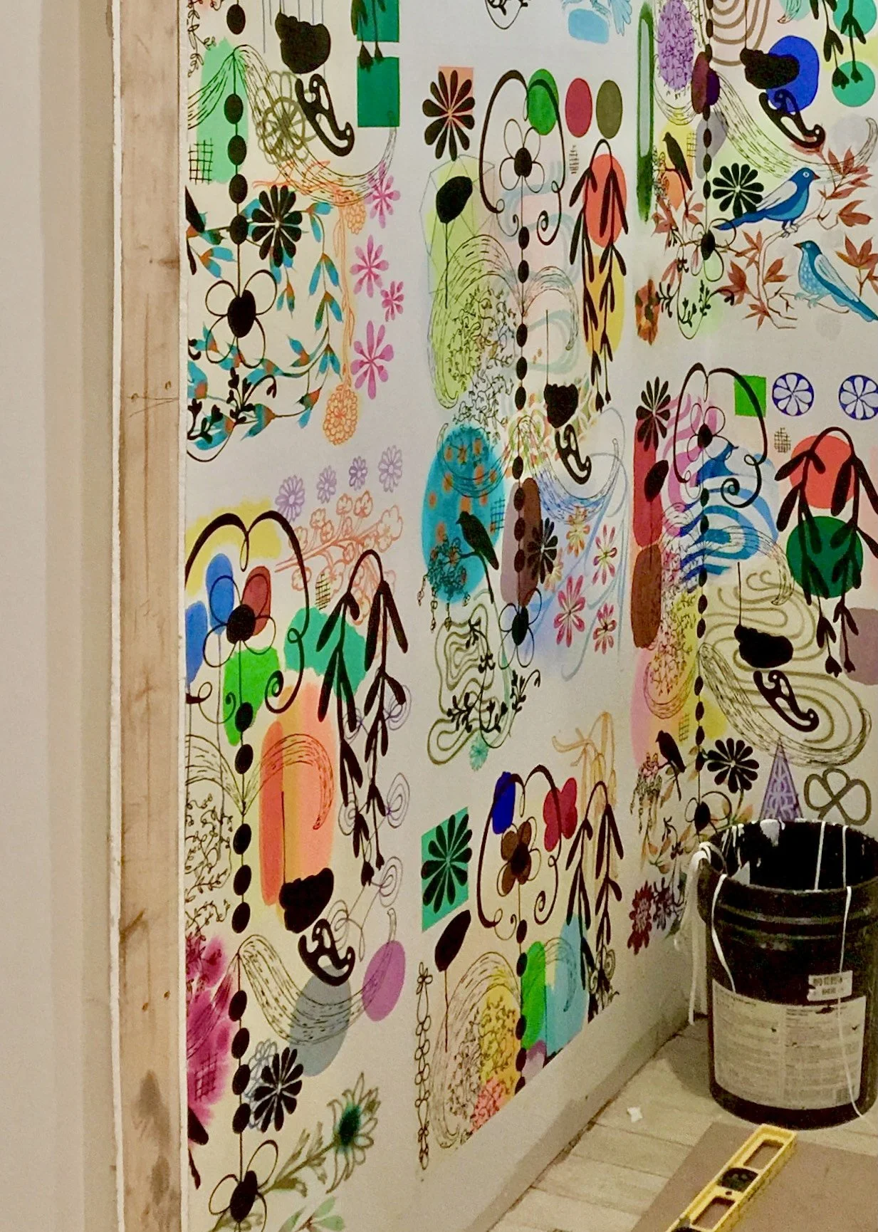 A colorful abstract painting with floral and animal motifs on a wall, partially surrounded by a wooden frame on the left and a black bucket on the bottom right.
