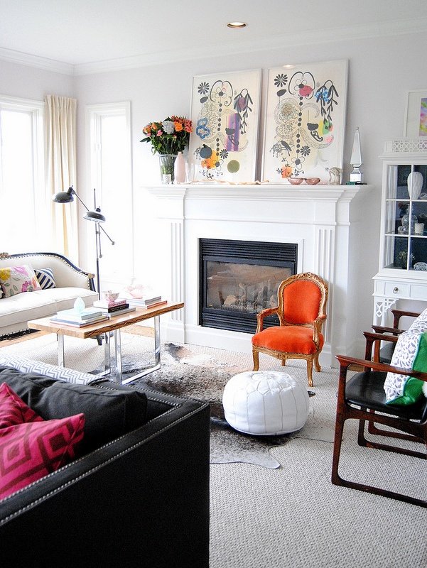 Living room with a white fireplace, colorful artwork above it, an orange velvet armchair, a white ottoman, a black sofa with pink pillows, a dining table with books, a white cabinet, and natural light from the windows.