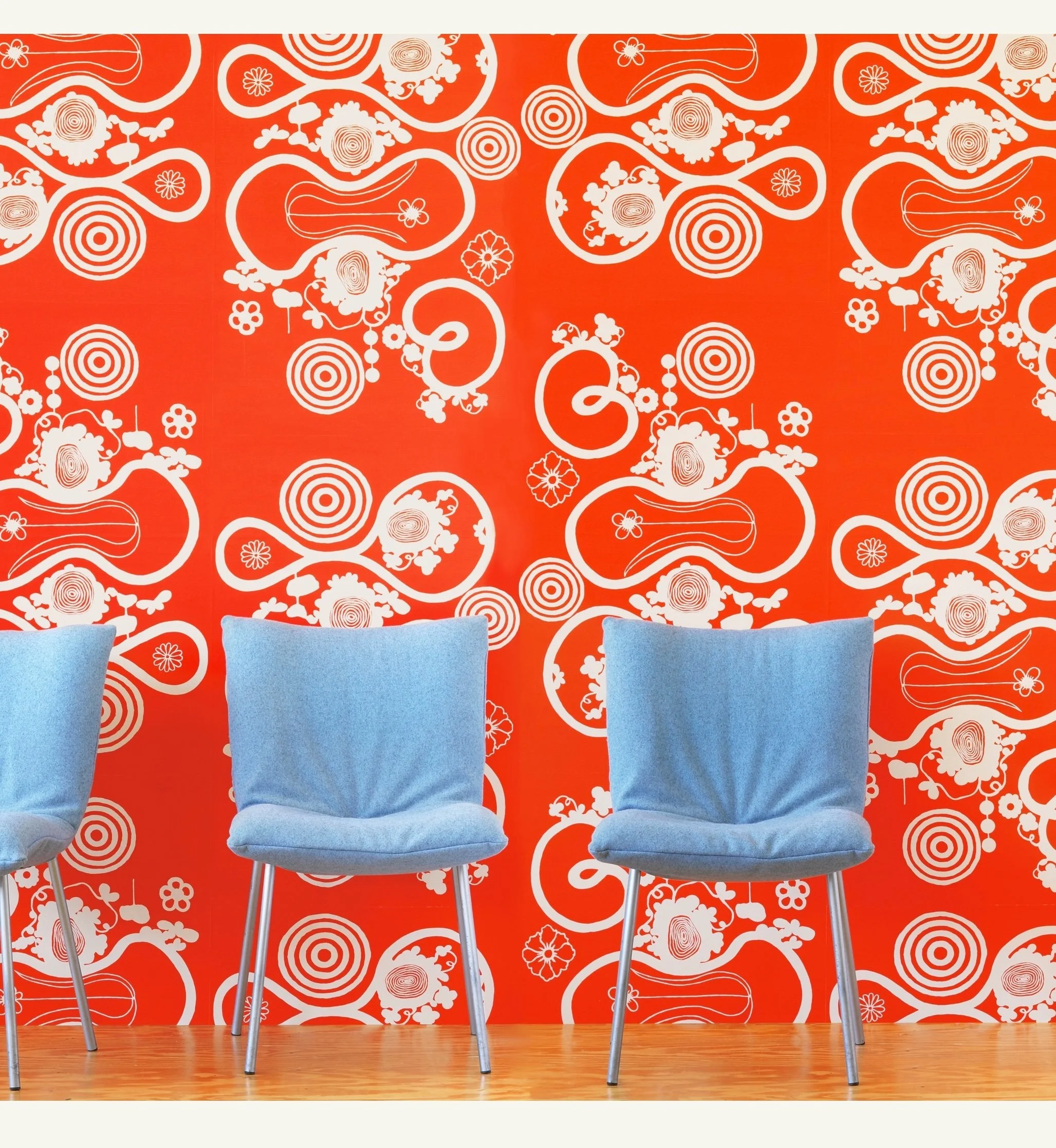 Three blue chairs with metal legs are placed against a vibrant red wall with a white abstract floral pattern. The floor is wooden.