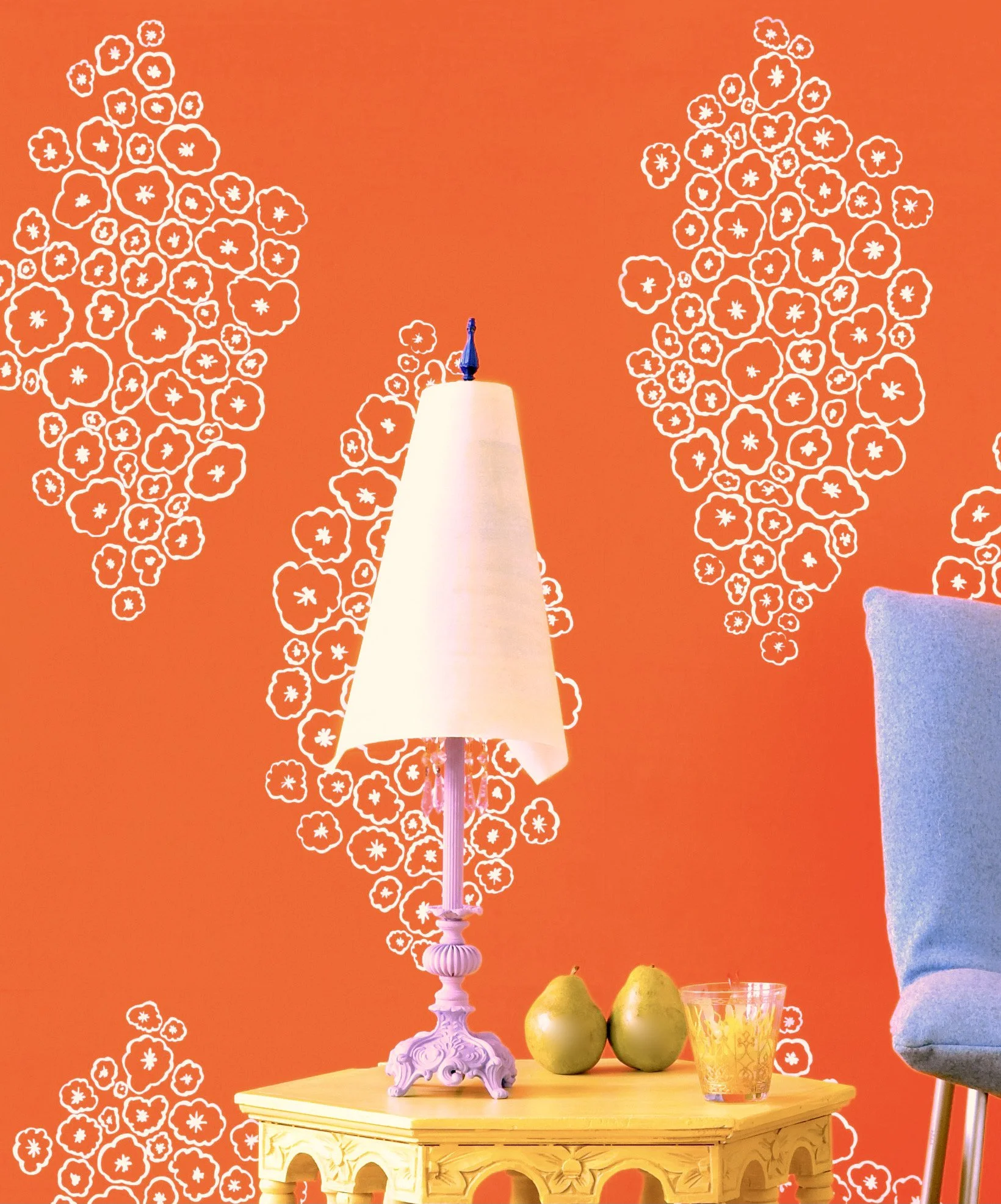 A yellow wooden table with a white ornate lamp, three green pears, a glass with an orange pattern, and a partial blue and white cloth on the right side, against an orange wall with white floral patterns.