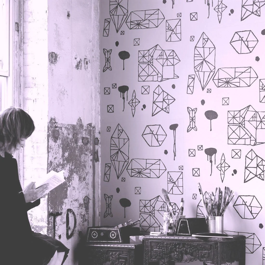 A woman sitting on a wooden chair, reading a book, in a room with a textured wall on the left and a patterned wall with geometric line art on the right. The desk below the patterned wall holds art supplies like brushes and containers.