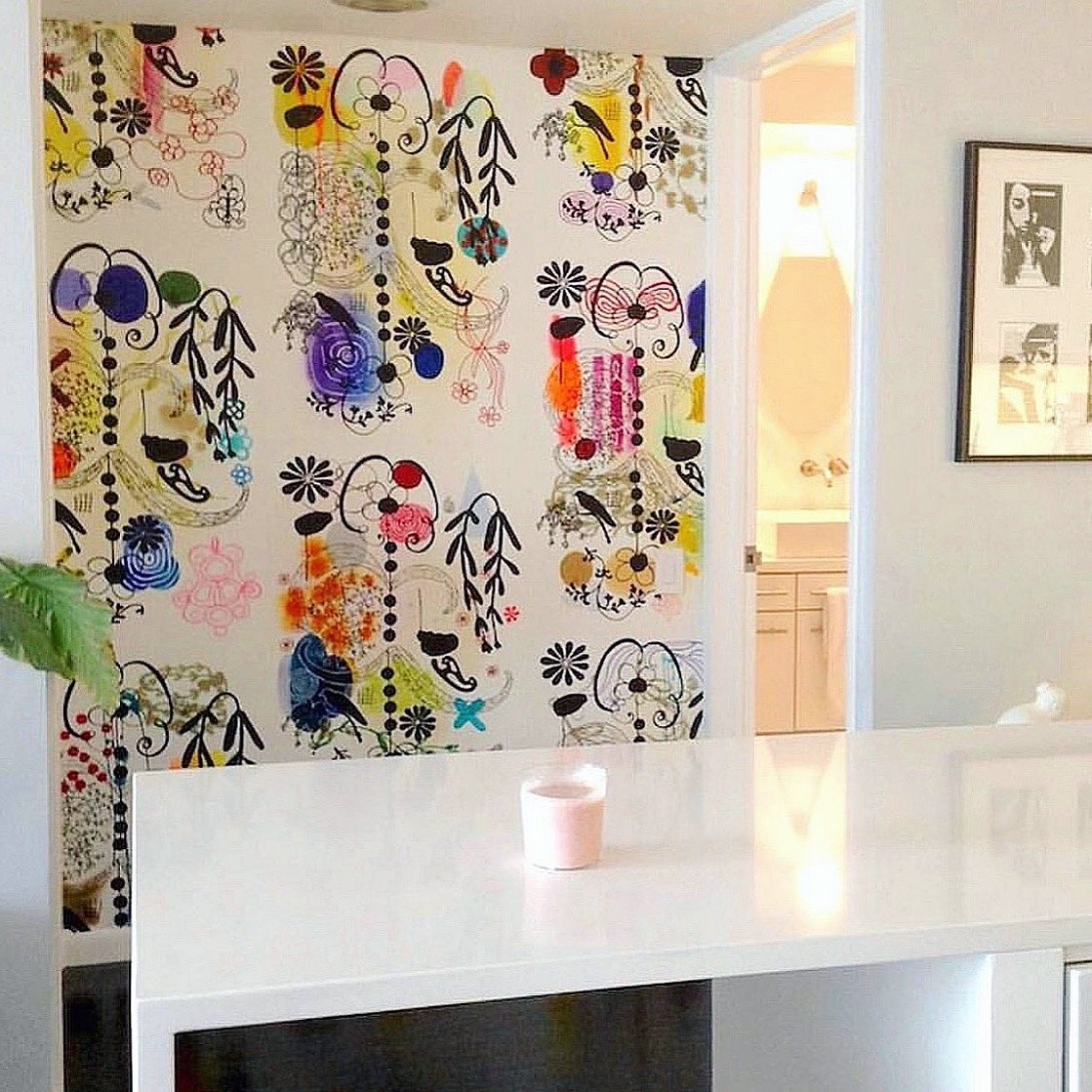 Colorful abstract patterned wall hanging in a bright room with a white counter, green plant, and framed black and white photos on the wall.