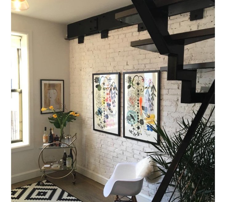 Interior of a modern living space with white brick wall, framed artwork, a white chair, a bar cart with bottles and glasses, a plant, and a black staircase.