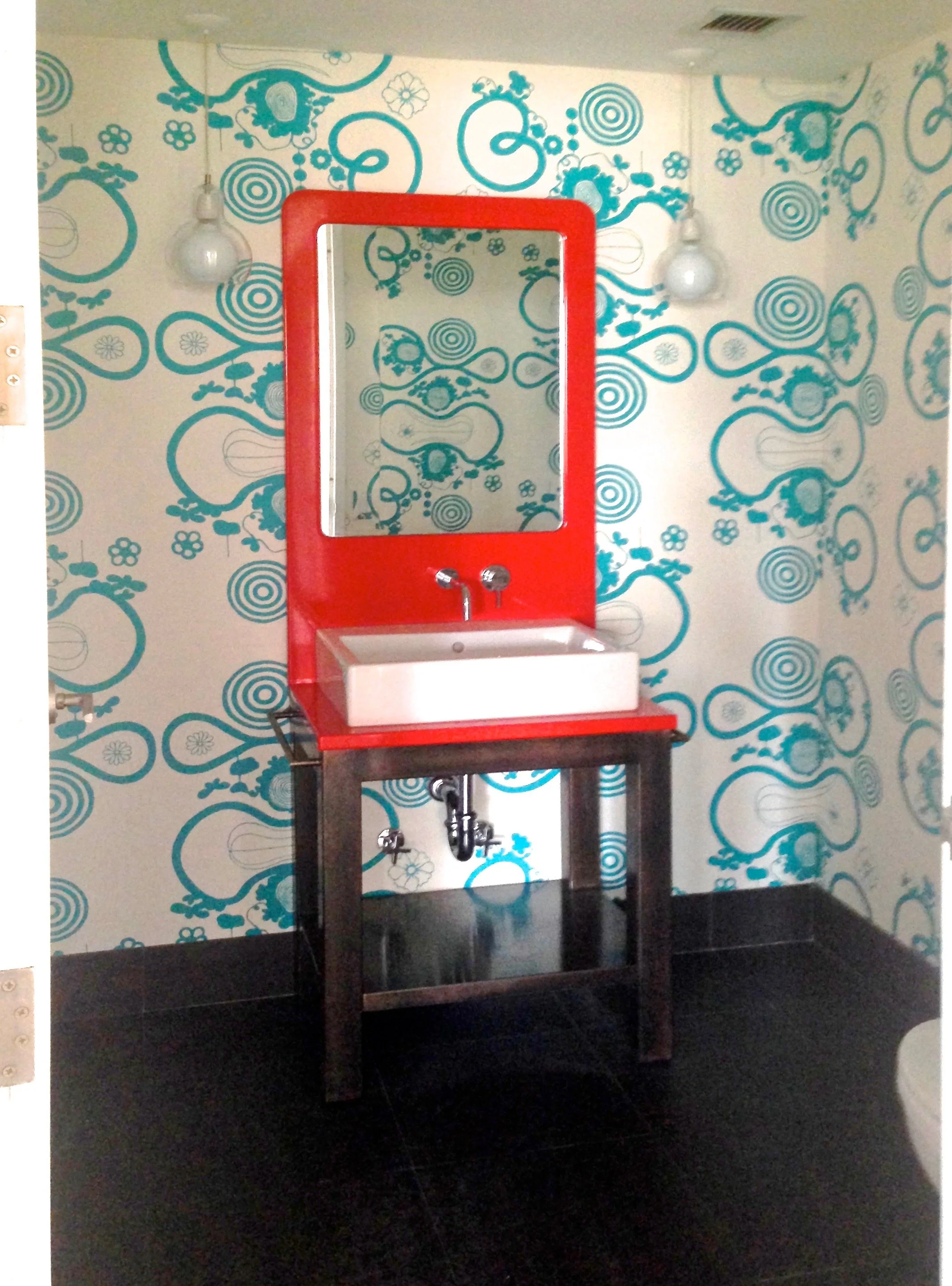 A bathroom with a red framed mirror above a small sink with faucet, set against a wall with blue floral and spiral pattern wallpaper. There are two white light fixtures above the mirror and a black stand beneath the sink.