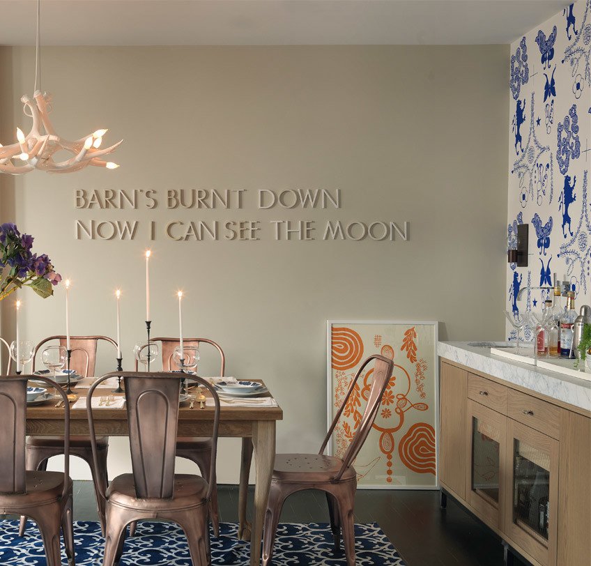 A dining room with a wooden table and four chairs, lit candles, a chandelier, a floral arrangement, and artwork on the wall with the text 'BARN'S BURNT DOWN NOW I CAN SEE THE MOON'. There are patterned wallpaper and a cabinet with a sink.