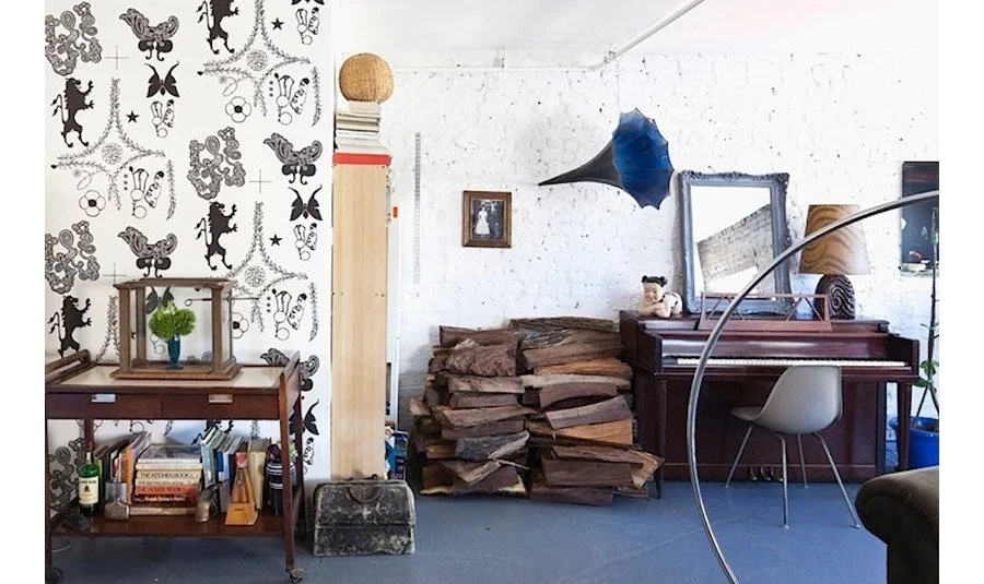 A cozy room with a white brick wall, decorated with a vintage mirror, a piano with a plush toy, and a stack of firewood. There is wallpaper with a playful animal and insect pattern on one wall, a small side table with books and a green plant, and a f