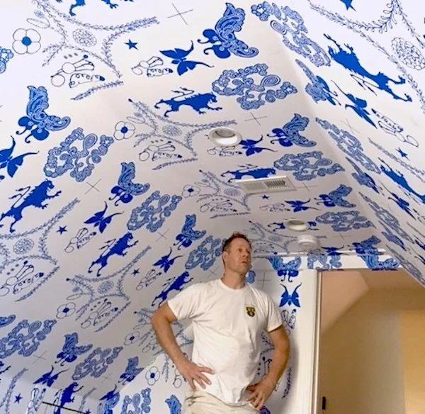 A man standing with hands on hips in a room with a ceiling and wall covered in blue and white patterned wallpaper with floral and butterfly designs.