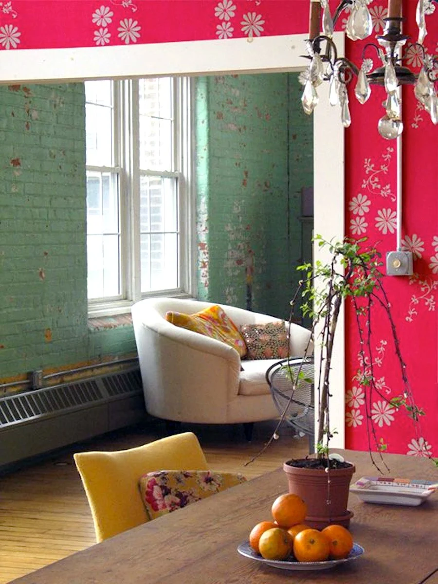 A cozy living room with green-painted brick walls, a white armchair with patterned pillows, a yellow chair with a floral pillow, and a wooden table with oranges and a potted plant. Bright natural light comes through large windows, and a decorative ch