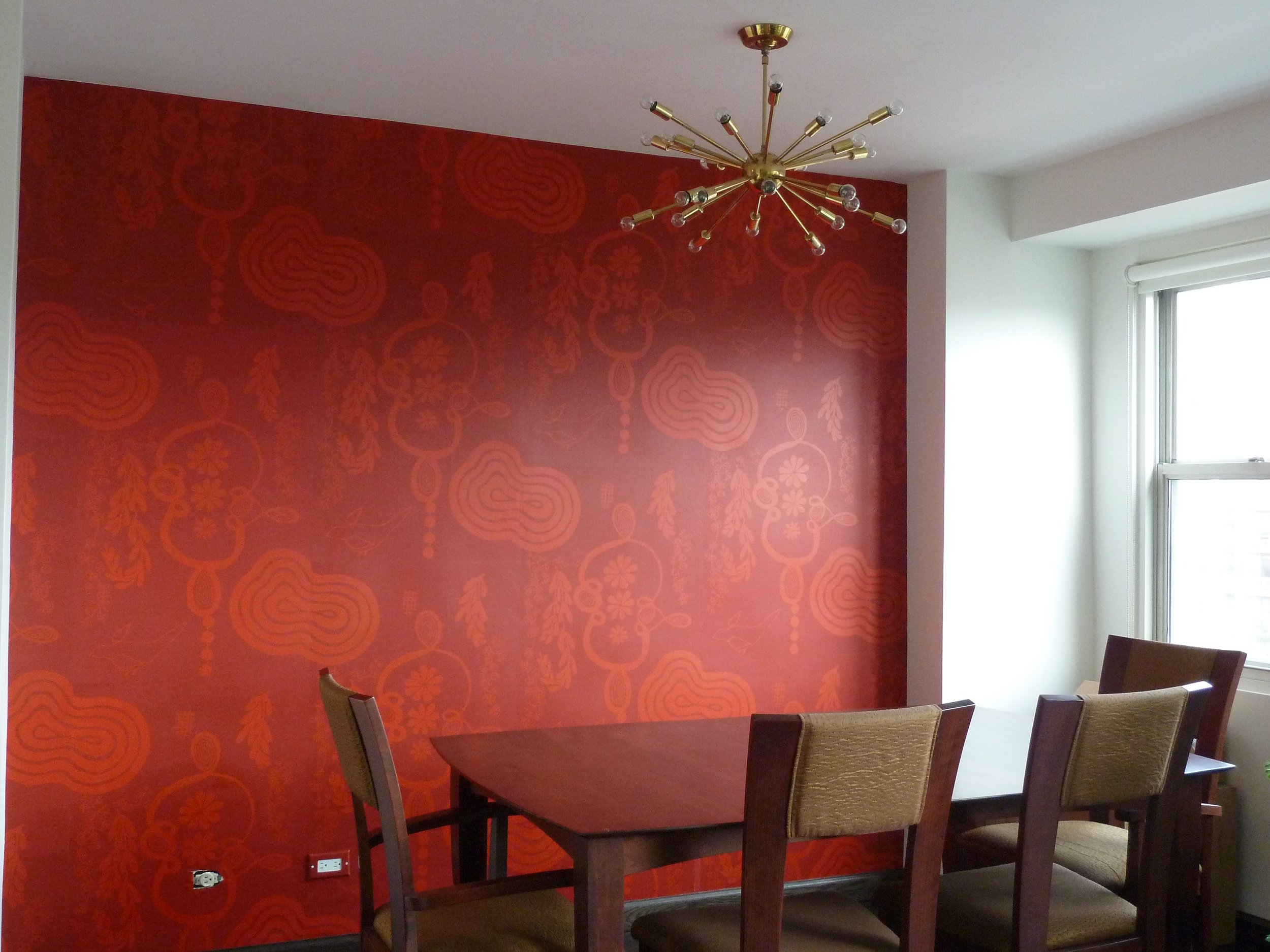 A dining room with a red wallpapered accent wall featuring a floral pattern, a modern gold chandelier hanging from the ceiling, a window with a white blind, a wooden dining table, and beige upholstered chairs.