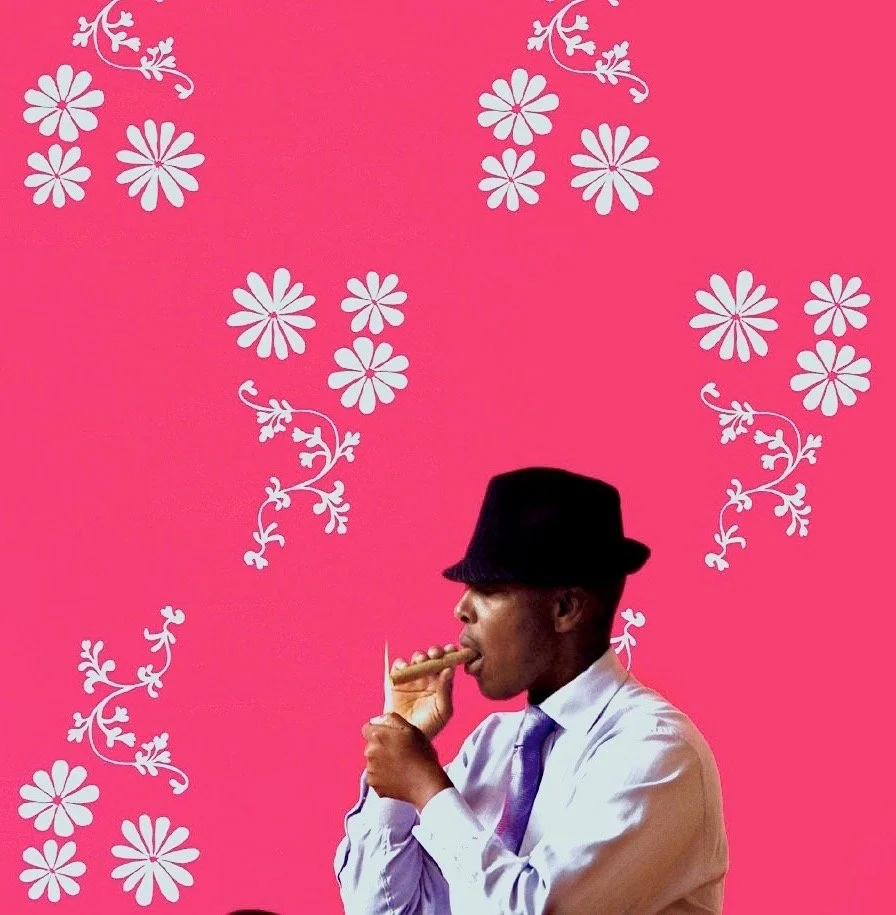 A man wearing a white shirt, purple tie, and black fedora hat, standing against a bright pink background with white floral patterns, smoking a cigar.