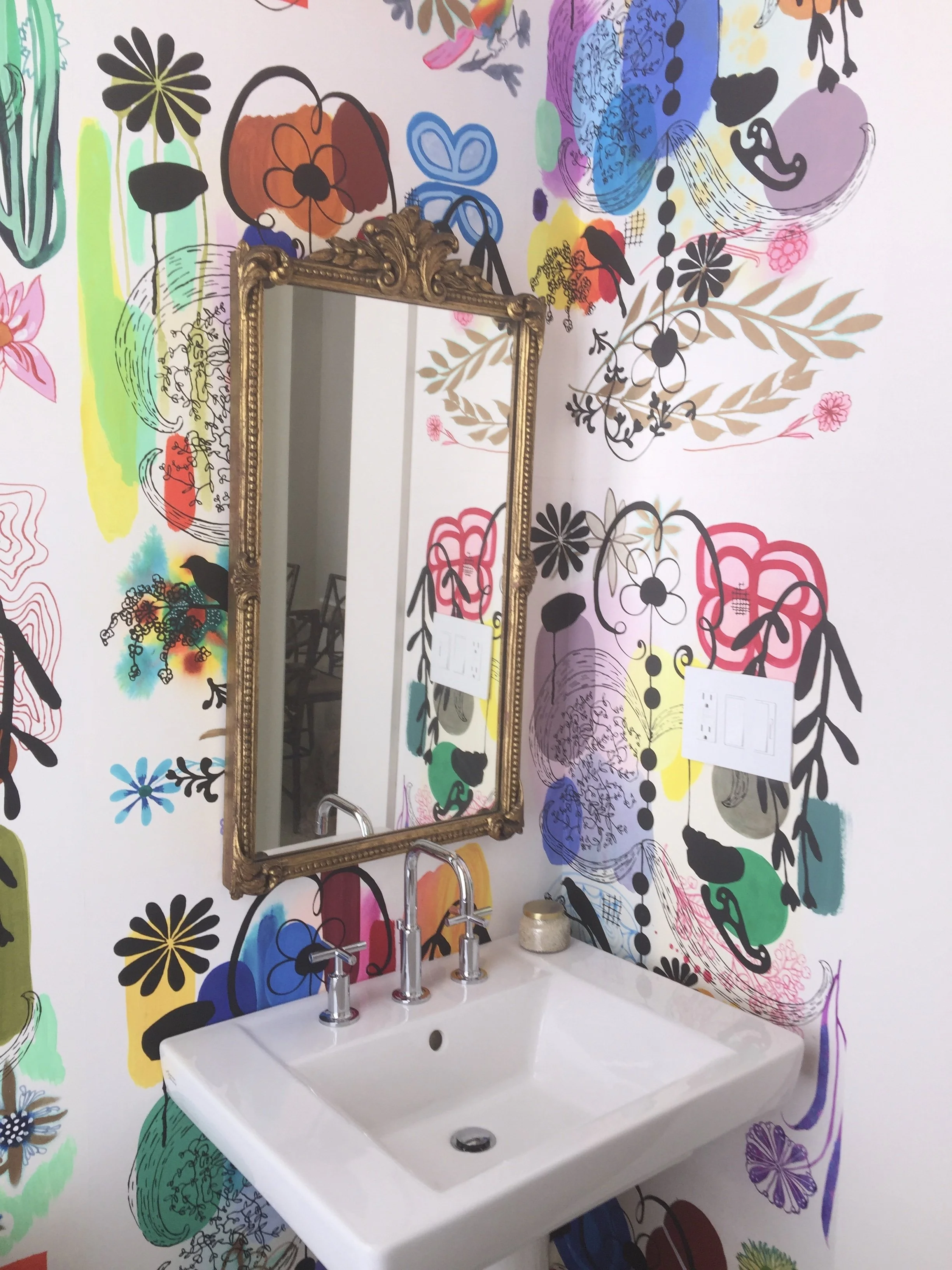 A white bathroom sink with a silver faucet mounted on a colorful, floral-patterned wall. The wall has various abstract and floral designs in multiple colors, including black, purple, pink, yellow, green, and blue. A gold ornate mirror hangs above the