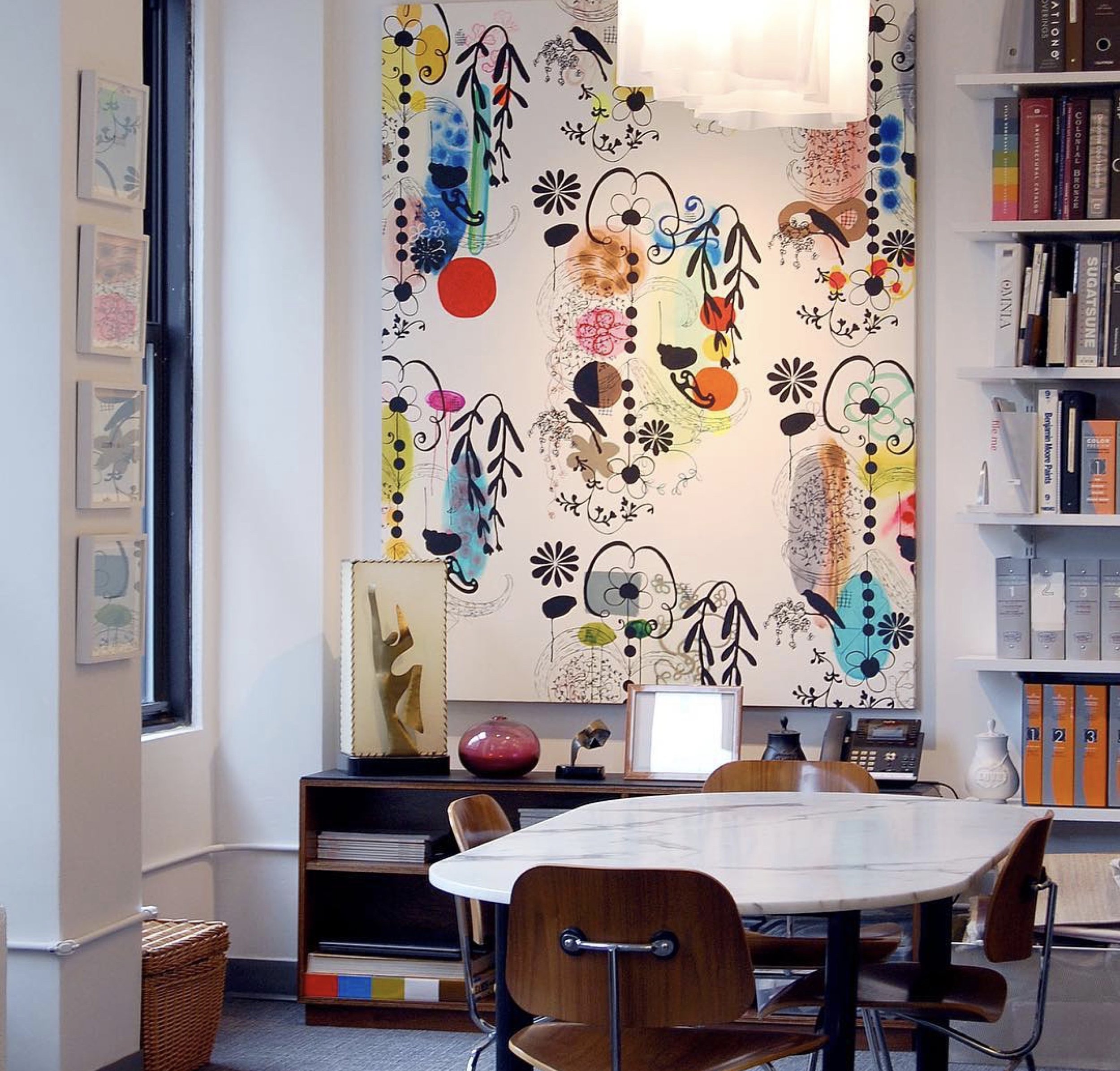 A cozy reading nook inside a home or office space. The room features a white wall decorated with whimsical, colorful floral and abstract artwork. A round white table with wooden chairs is set in the foreground. To the right, there is a white bookshel