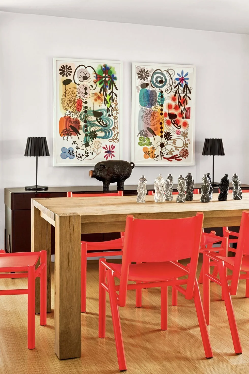 Modern dining room with a wooden table, red chairs, black lamps, colorful abstract wall art, and decorative objects including a pig sculpture and glass bottles.