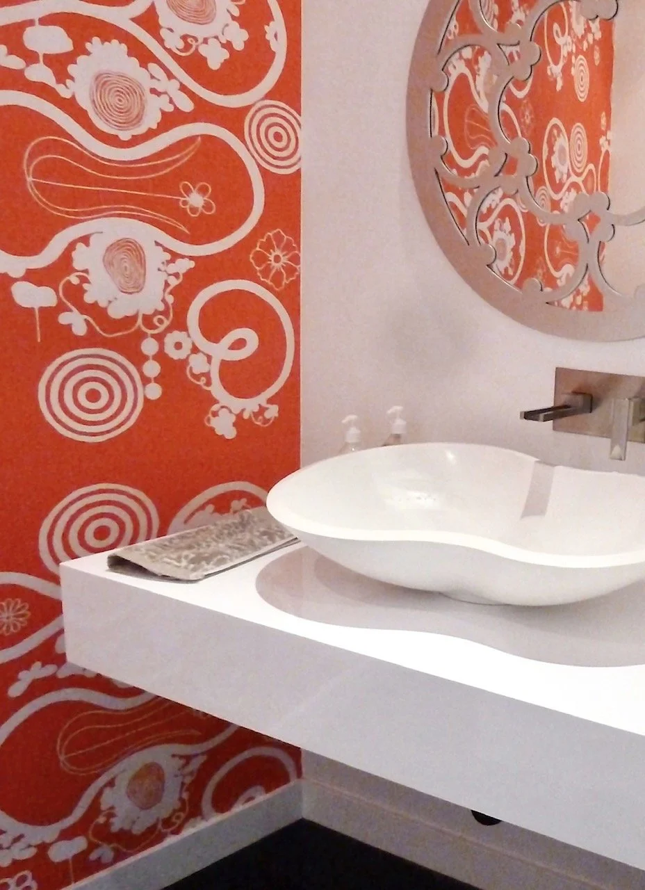 Bathroom with a white vessel sink on a white countertop, a silver decorative wall piece, a patterned towel, red and white abstract wallpaper, and two small soap dispensers.