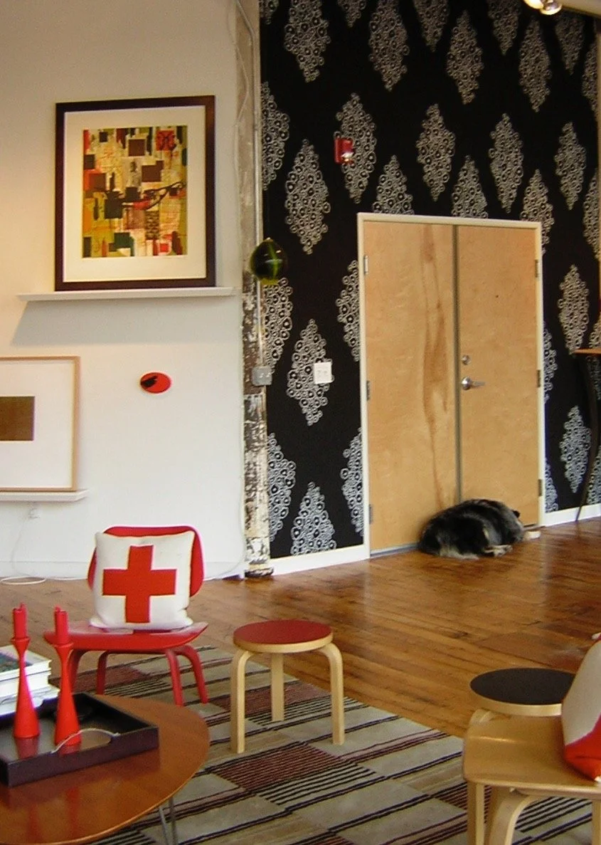 Living room with Black and white damask wallpaper, a closed wooden door, a black and brown dog sleeping on the floor, a table with red candles, framed artwork on the wall, and a red and white Red Cross bag on a chair.
