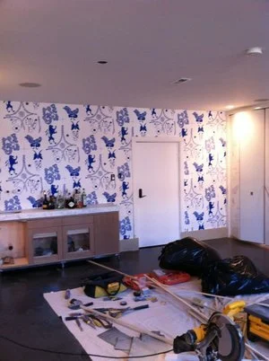 Room under renovation with construction tools, black bag, and work materials on the floor, and a blue and white cartoon-themed wallpaper on the wall.