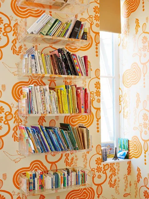 A set of transparent wall-mounted bookshelves filled with books in a room with orange and white patterned wallpaper, next to a window with a view of the outdoors.