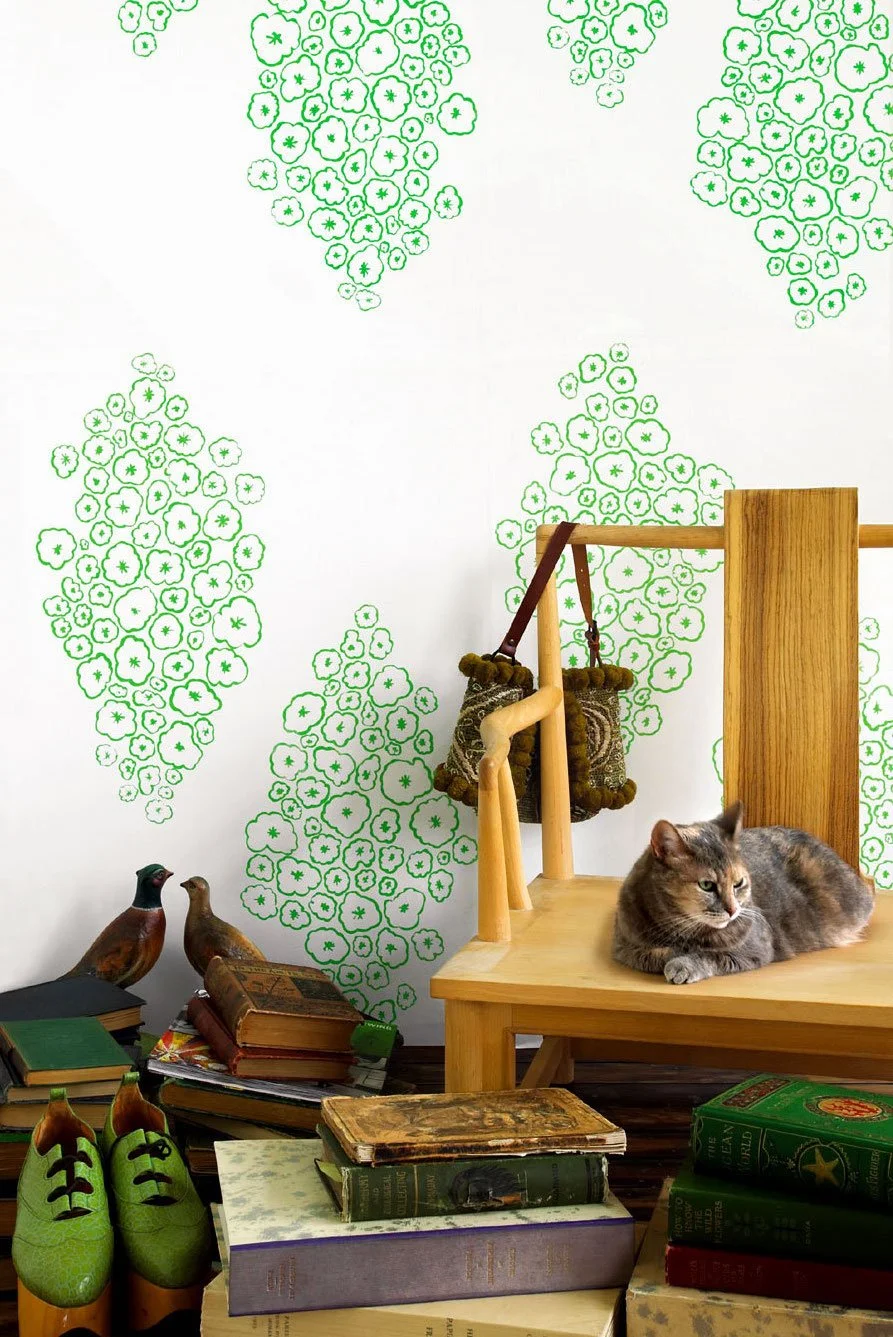 A cozy room corner with a wooden chair, a gray tabby cat resting on it, a wall decorated with green floral pattern wallpaper, a pair of wooden bird figurines, a stack of books on the floor, and a pair of green slippers.