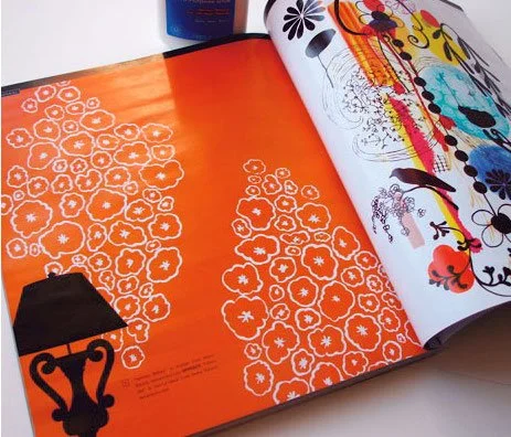 Open magazine showing two pages with floral and colorful abstract designs, and an orange background with white floral patterns.
