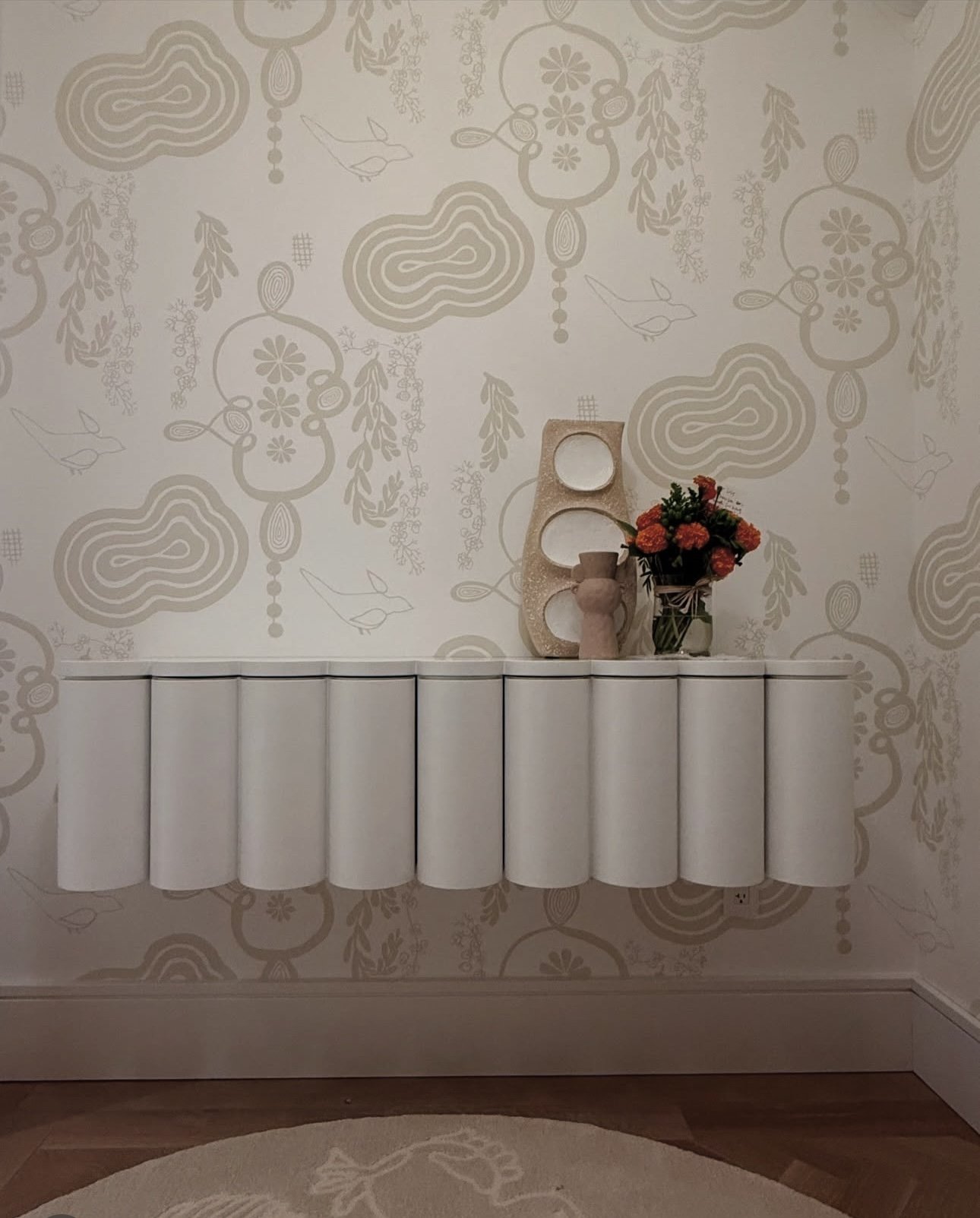 A white decorative wall with beige abstract patterns, featuring line drawings of birds and floral motifs. A white radiator extends horizontally beneath a small arrangement of orange flowers in a glass vase, a beige sculptural piece, and a small beige