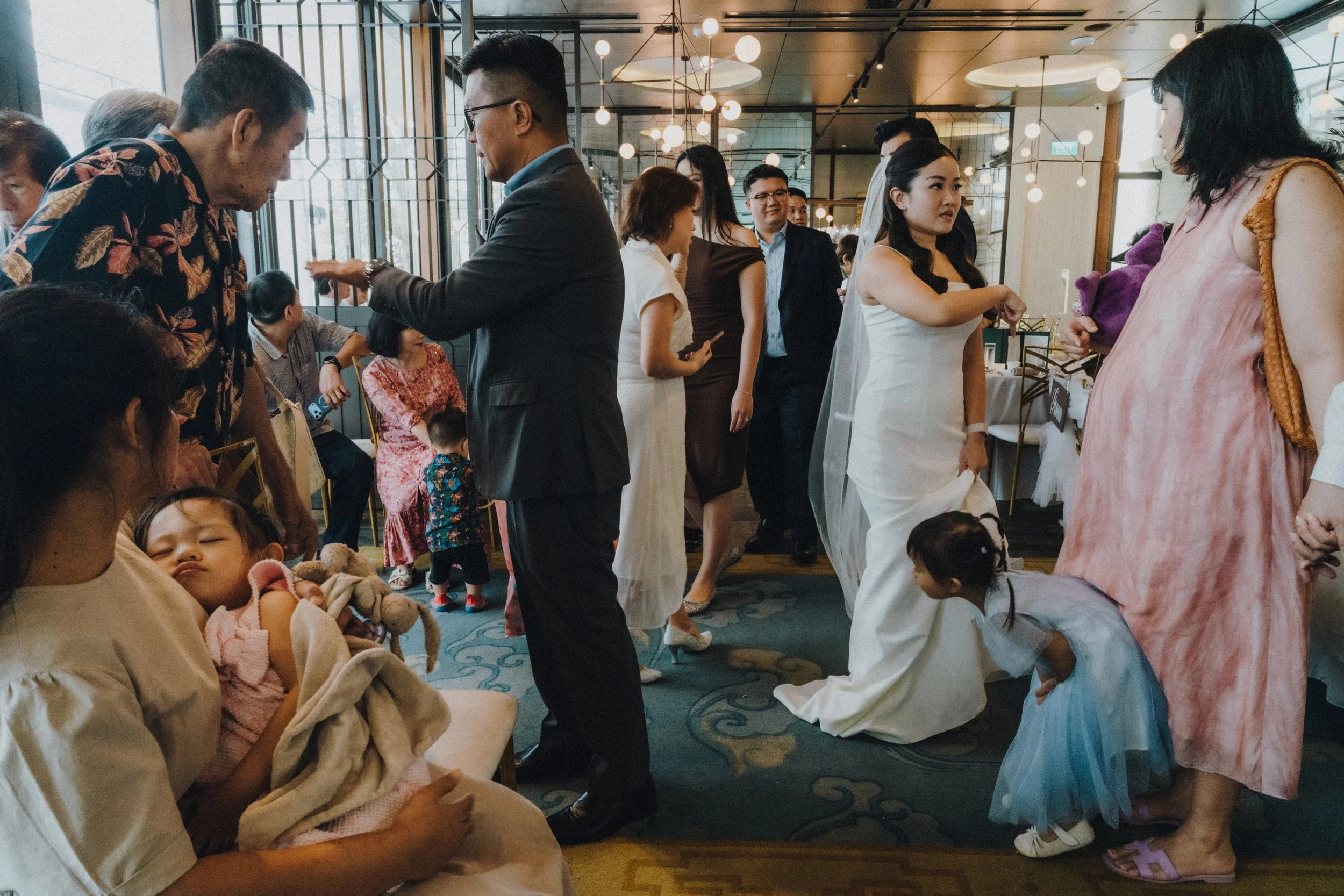 A diverse group of people, including children and adults, gathered indoors at a wedding, some engaged in conversation while others are seated. Authentic, unfiltered moments captured at wedding.