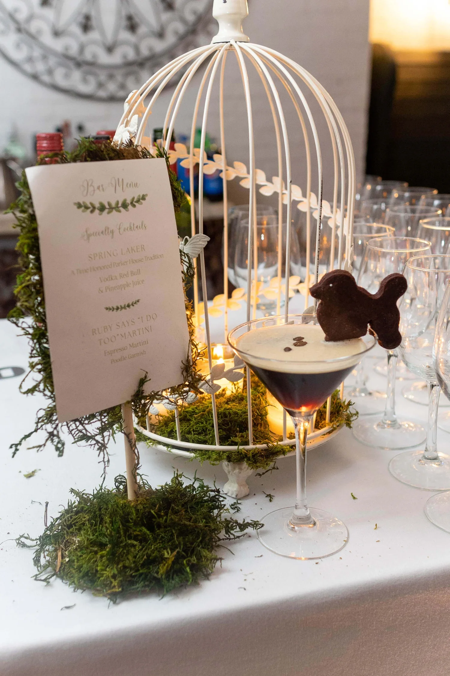 Styled wedding bar setup featuring a specialty cocktail sign, glassware, candles, and an espresso martini with a dog-shaped garnish.