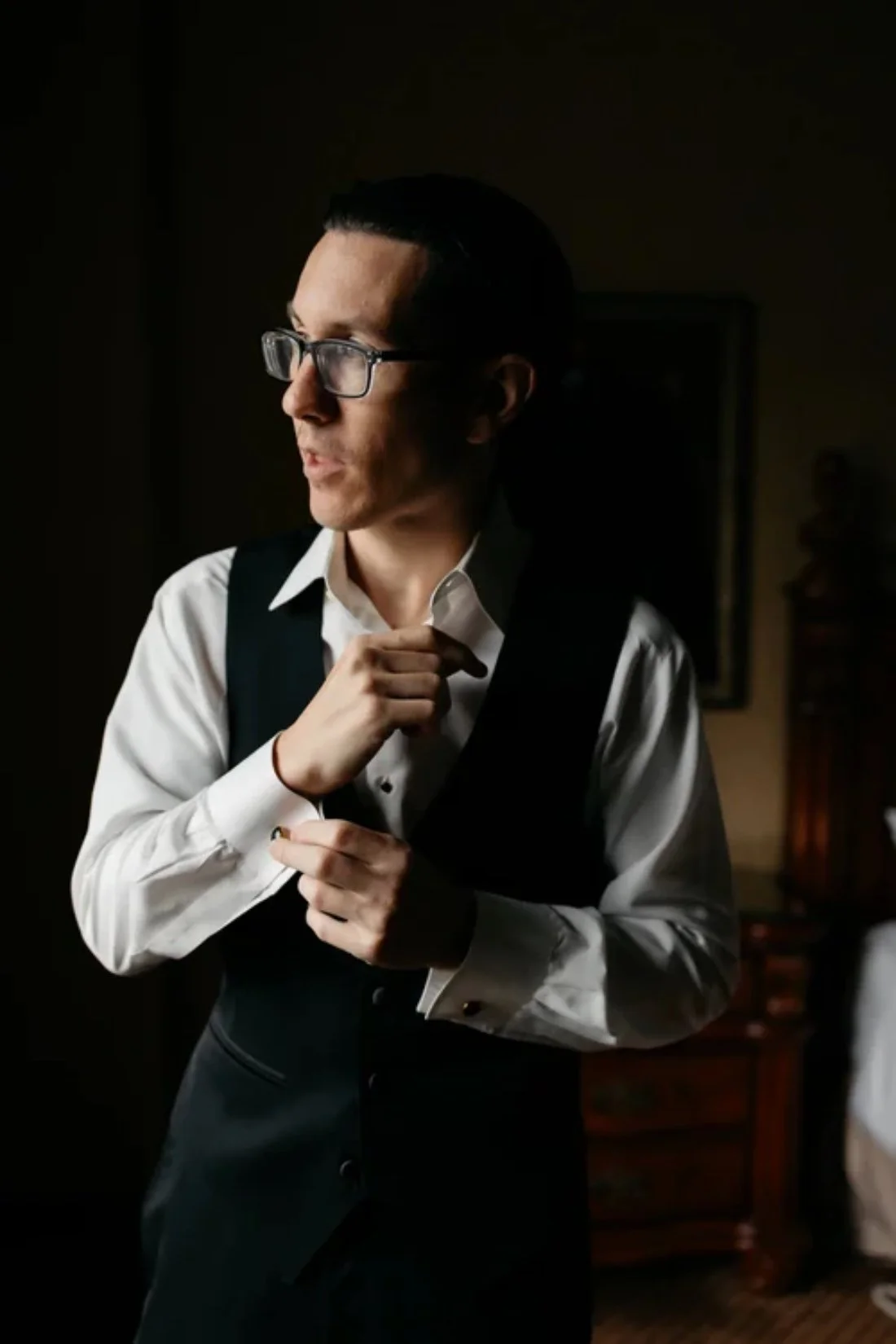 A groom adjusting his cufflinks in a dimly lit room, wearing a white shirt and dark vest that reflect classic winter wedding fashion for men.