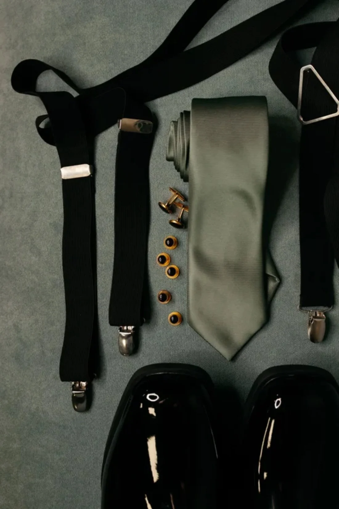 A flat lay of groom accessories including suspenders, a sage green tie, black shoes, and gold cufflinks, highlighting refined winter wedding fashion details for grooms.