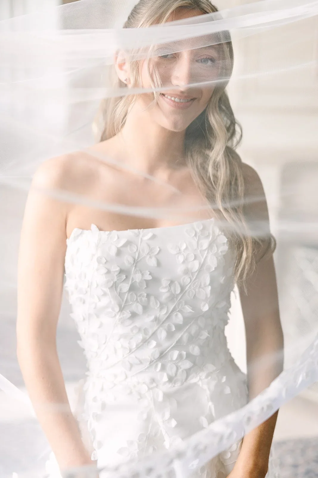 Bride posing softly behind her veil in natural light, showcasing delicate texture and timeless bridal details