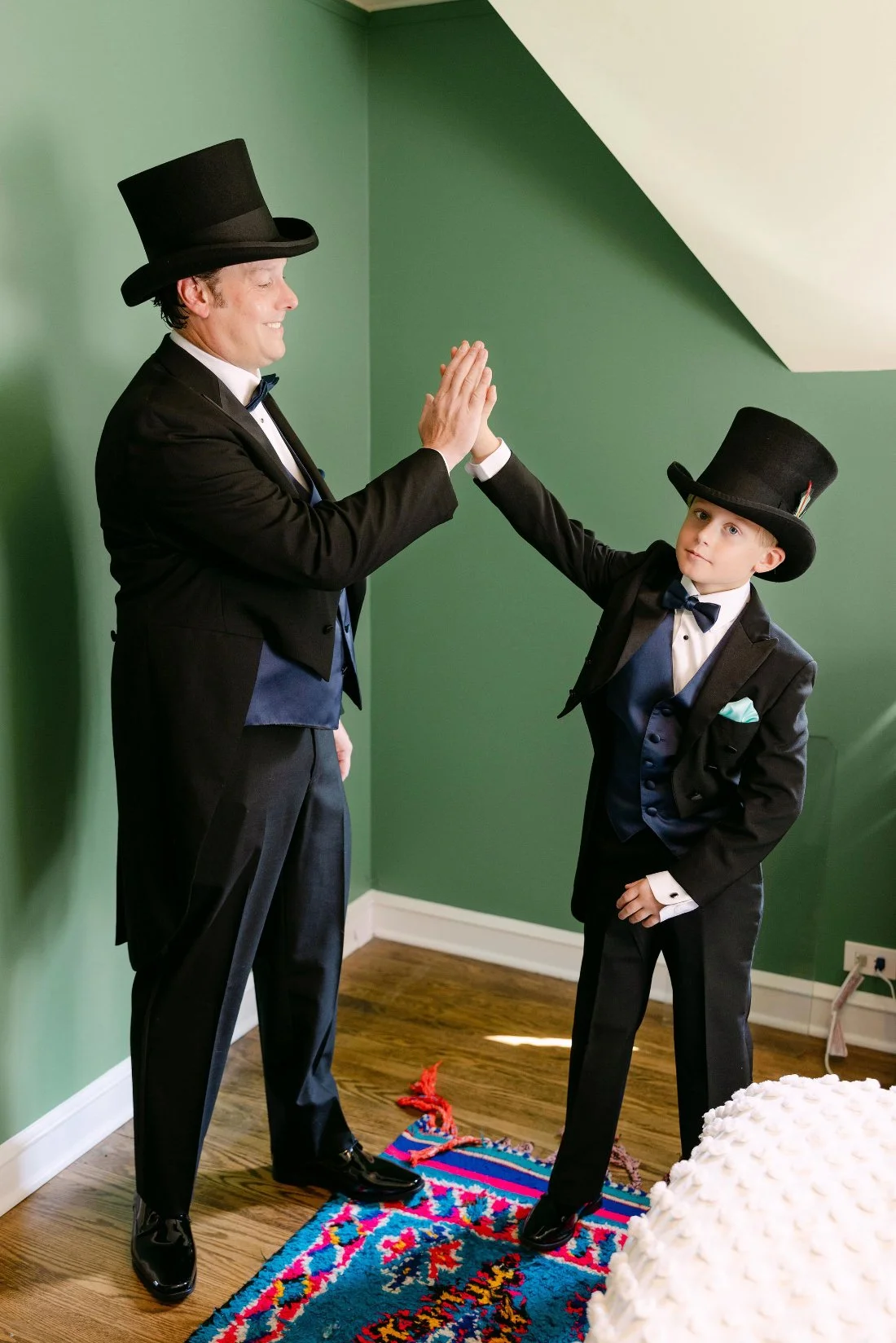 Groom and ring bearer sharing high fives in matching tuxedos and top hats before outdoor wedding in New York
